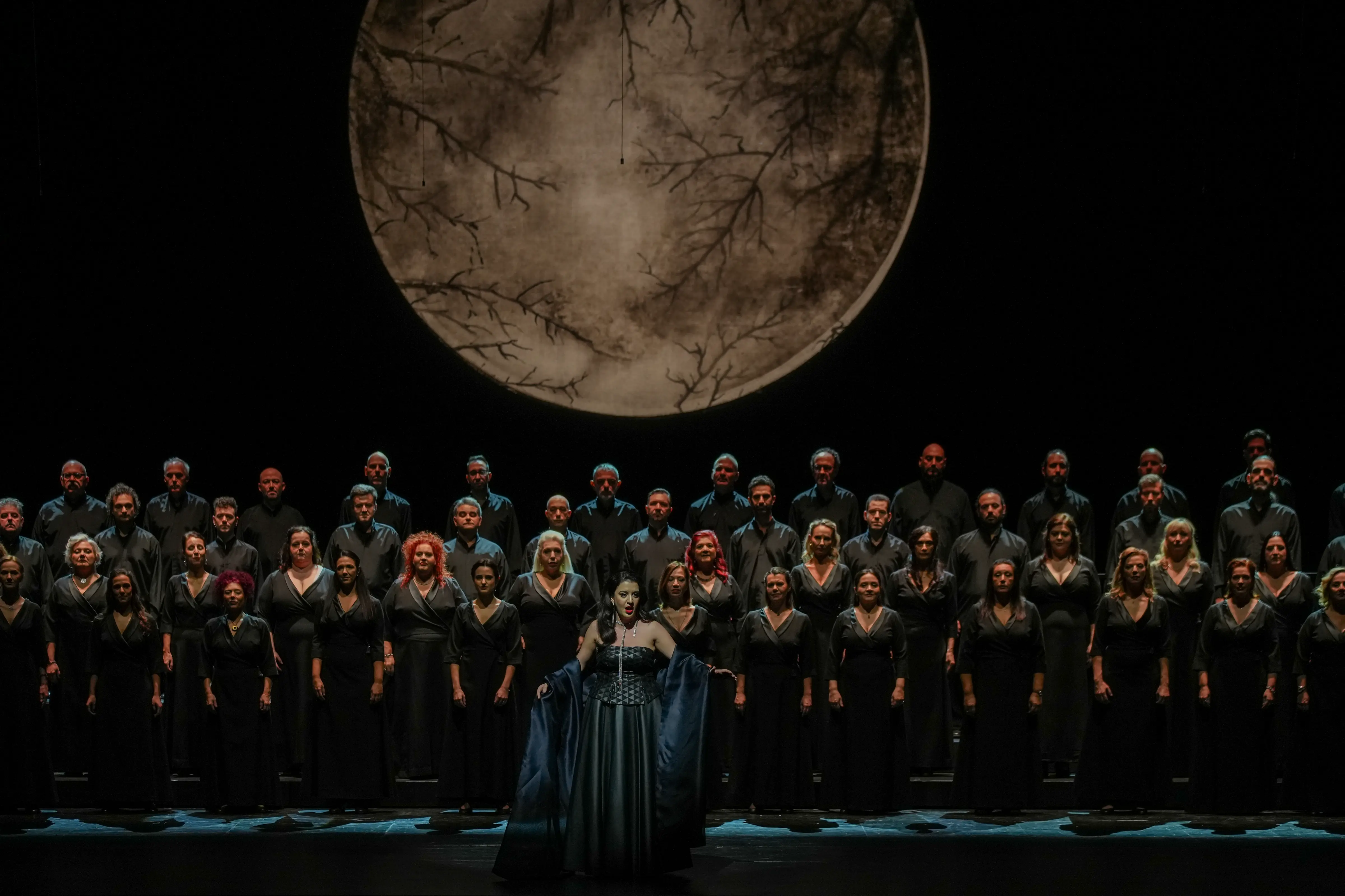 A solo opera singer performs in front of the Greek National Opera Chorus, standing beneath a dramatic full-moon backdrop during the 2025 International Opera Awards in Athens.