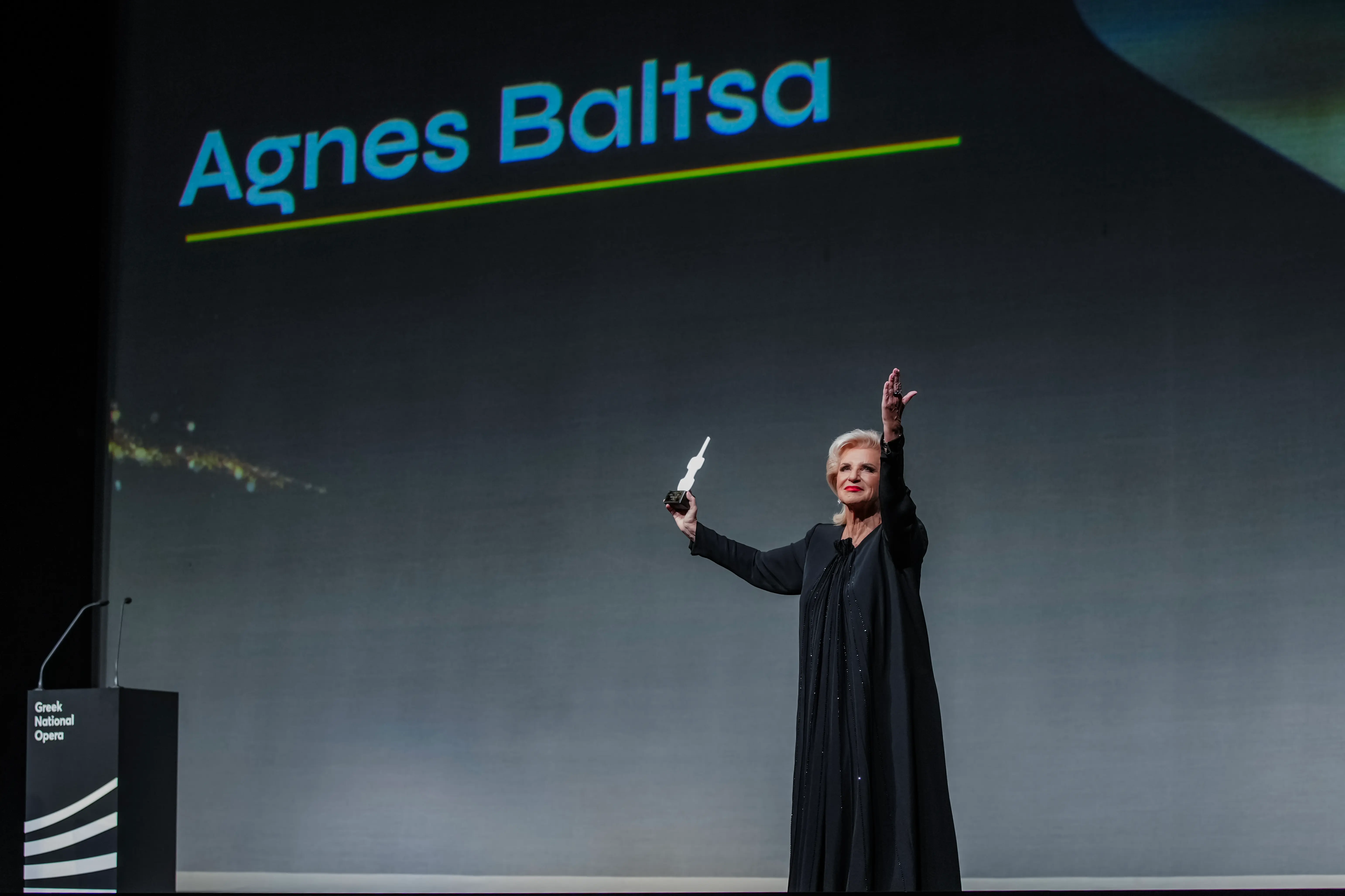 Agnes Baltsa stands on stage holding her Lifetime Achievement Award at the 2025 International Opera Awards, raising one arm in gratitude as her name is displayed in large letters behind her.