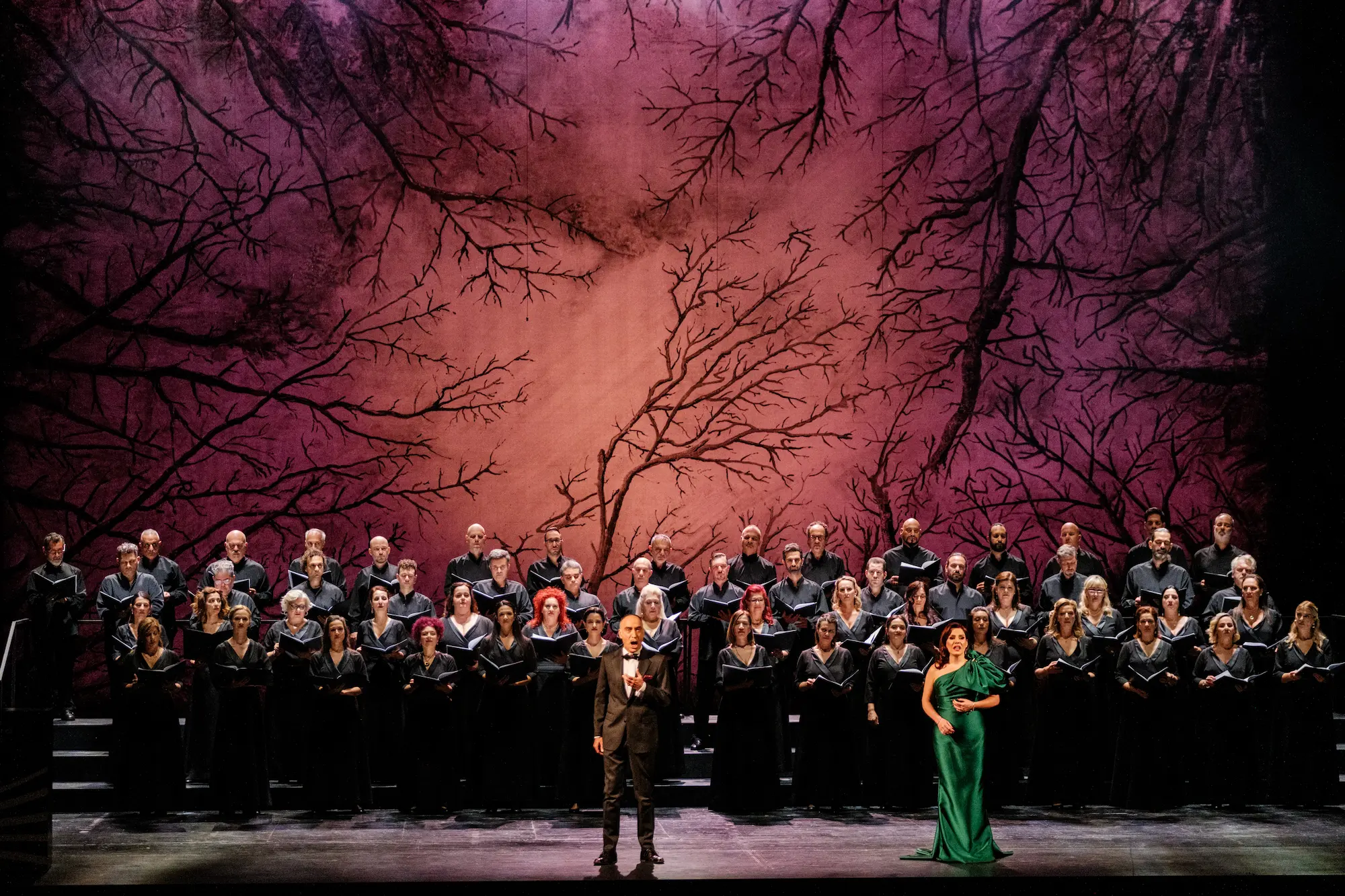 Yannis Christopoulos and Vassiliki Karayanni perform in front of the Greek National Opera Chorus, standing on a dramatic stage with a vivid red backdrop painted with dark, branching tree silhouettes.