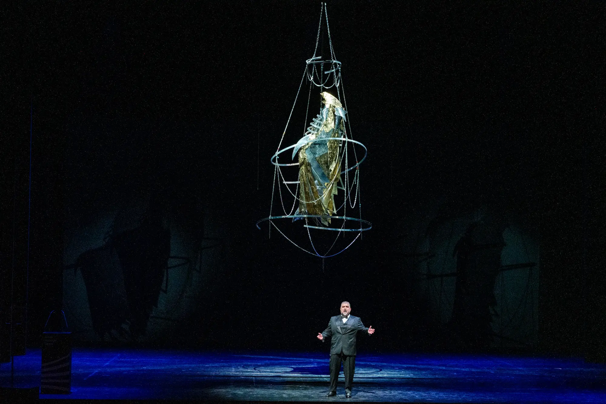 Dimitri Platanias performs on a dimly lit stage beneath a large, suspended metallic sculpture draped in gold, creating a dramatic and atmospheric operatic scene.