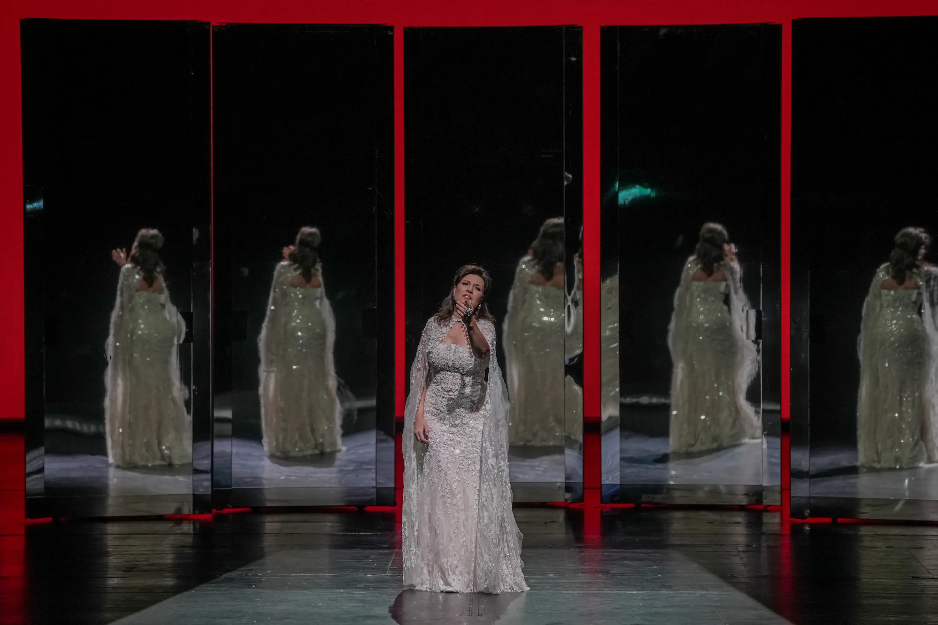 Marina Rebeka performs on stage in a shimmering white gown, standing before a row of tall mirrors framed in red, her reflections creating a dramatic and elegant visual effect.