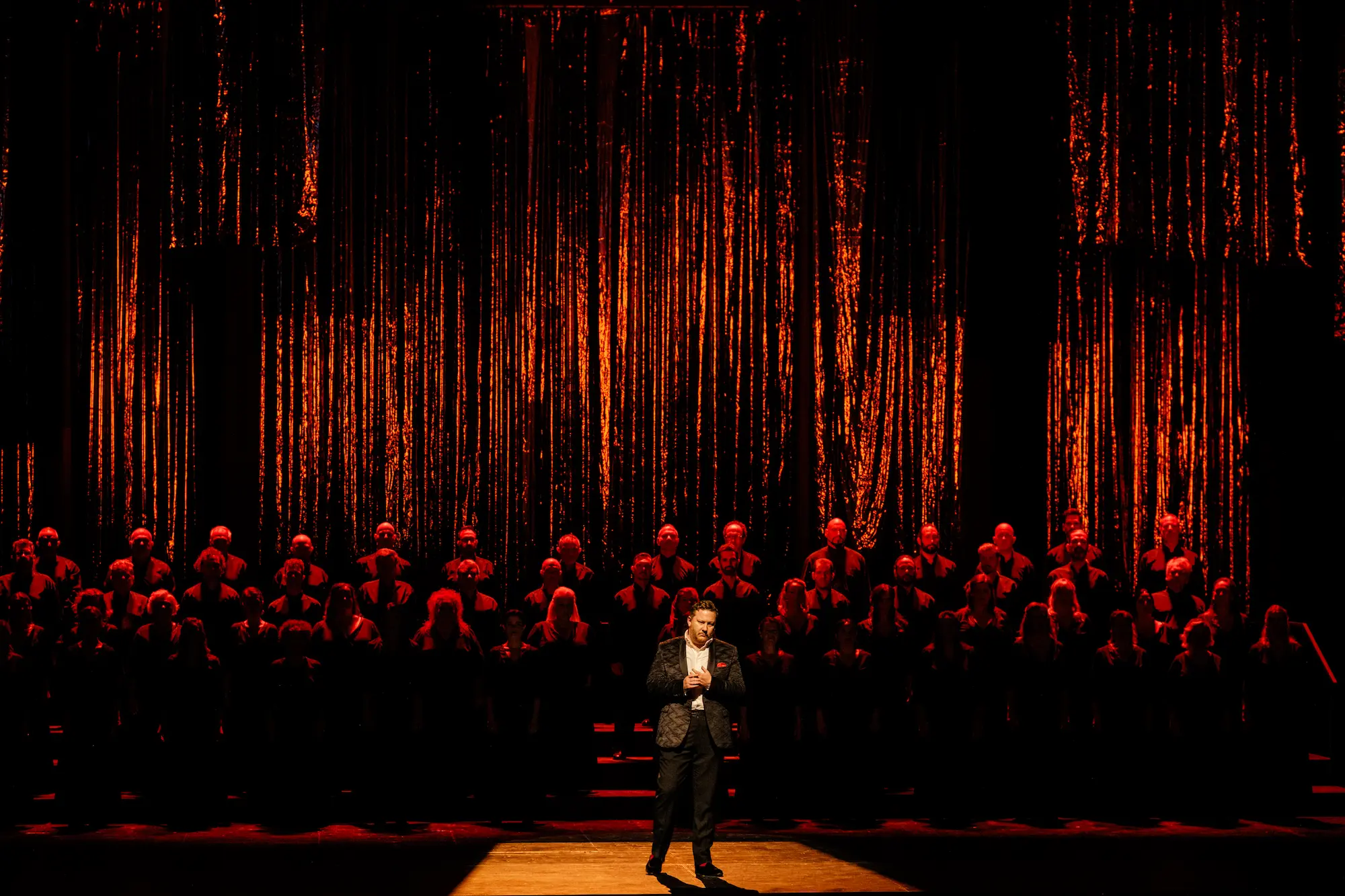 American bass-baritone Nicholas Brownlee performs on stage under dramatic red and gold lighting, standing before the Greek National Opera chorus illuminated in deep red tones.