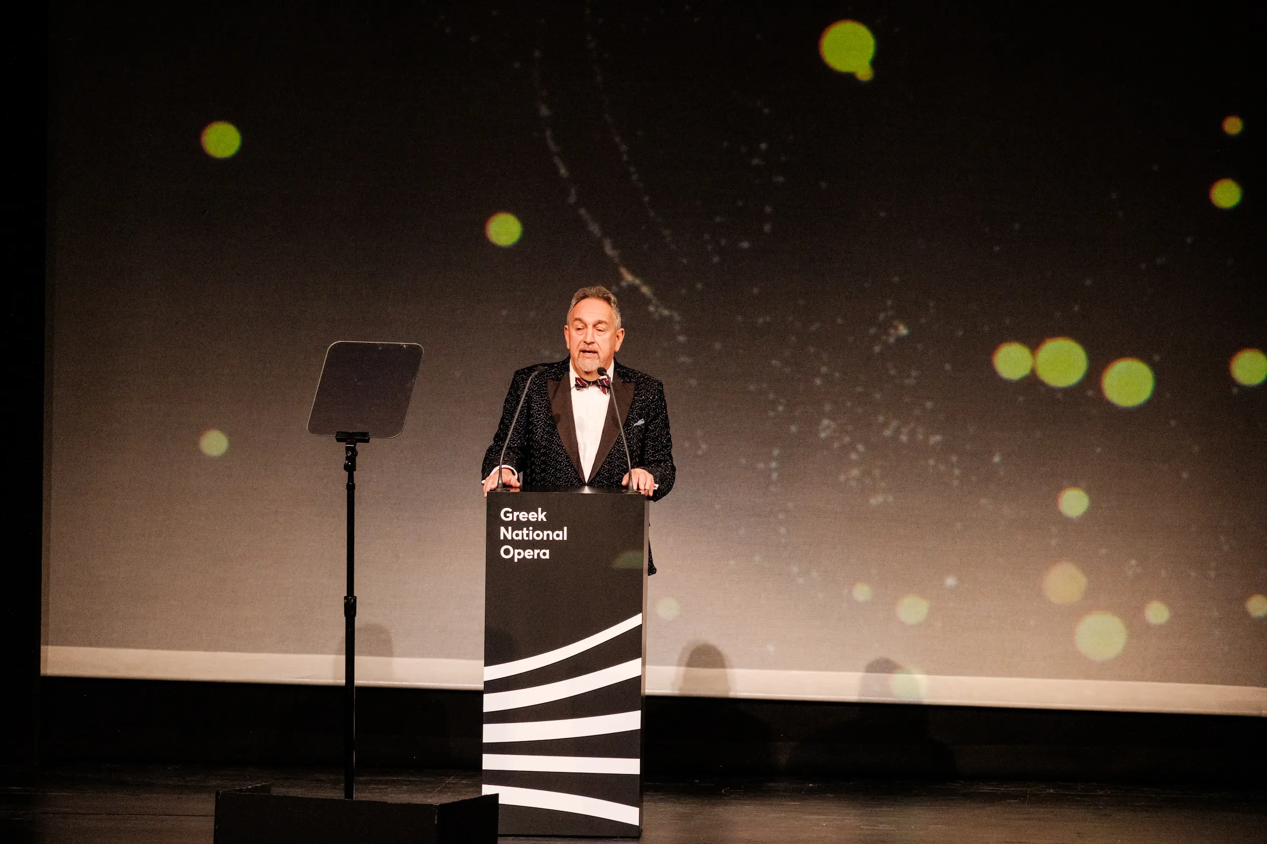 Harry Hyman, founder of the International Opera Awards, speaks at a podium marked “Greek National Opera” during the 2025 ceremony, standing before a dark stage backdrop with glowing light effects.