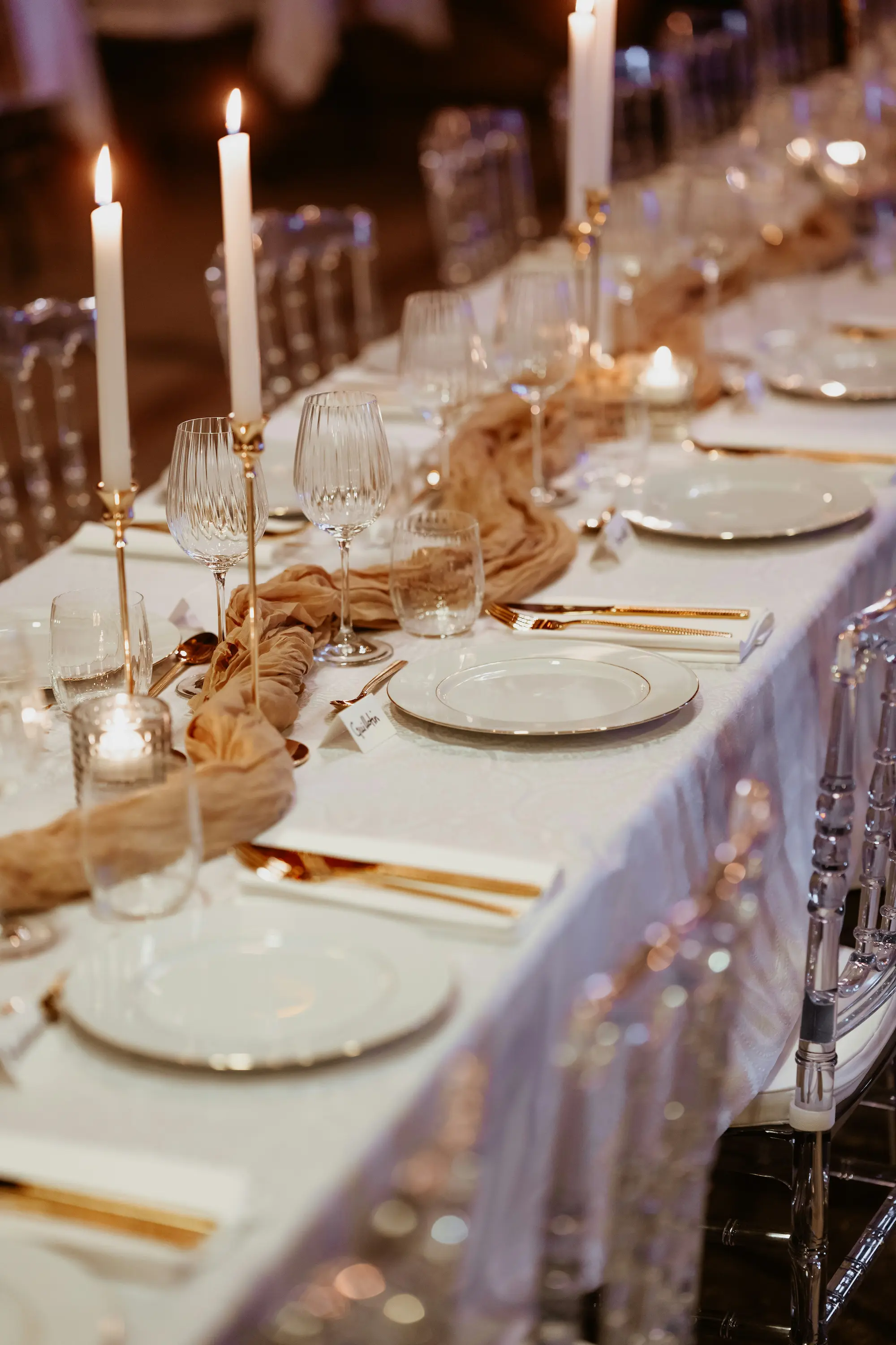 A refined, candlelit dinner table arranged with gold cutlery, crystal glassware, white plates, and soft fabric runners, creating a warm and luxurious atmosphere.