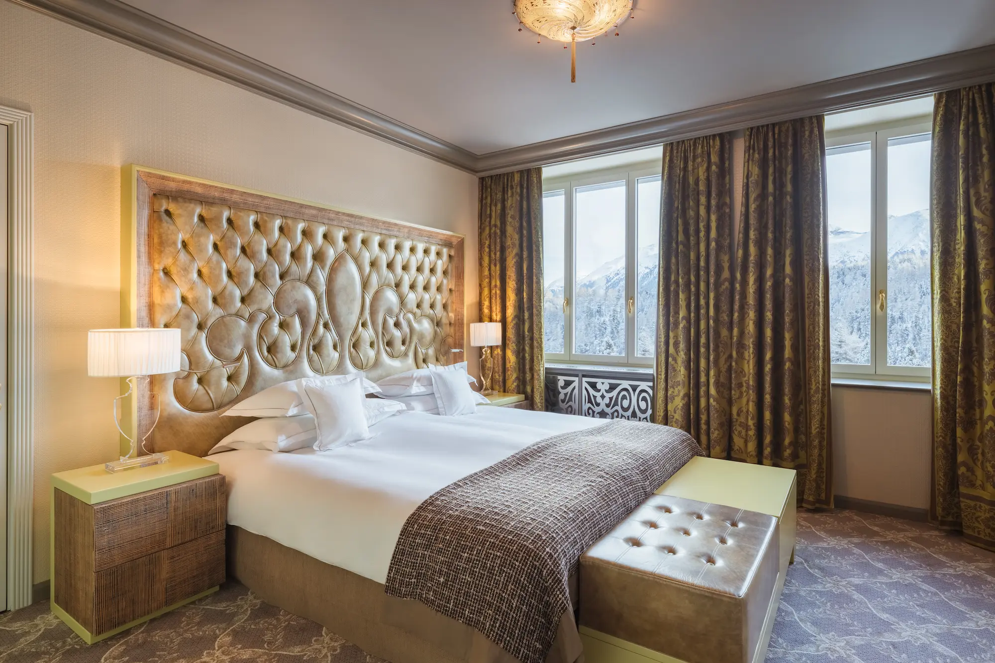 Elegant Carlton Hotel bedroom with ornate tufted leather headboard, crisp white linens, and floor-to-ceiling windows framing snow-covered alpine peaks