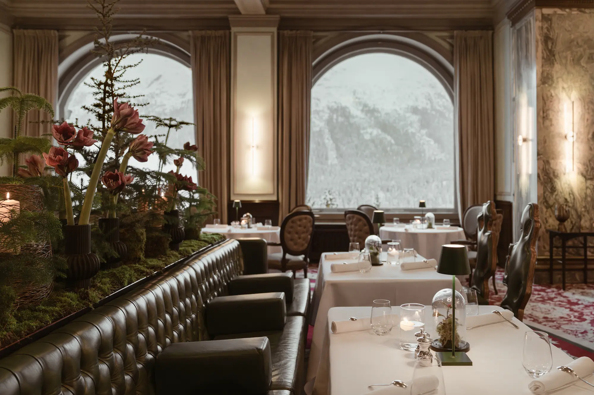 Elegant dining room at Carlton Hotel St. Moritz with tufted leather banquettes, white tablecloths, and arched windows overlooking snowy mountains
