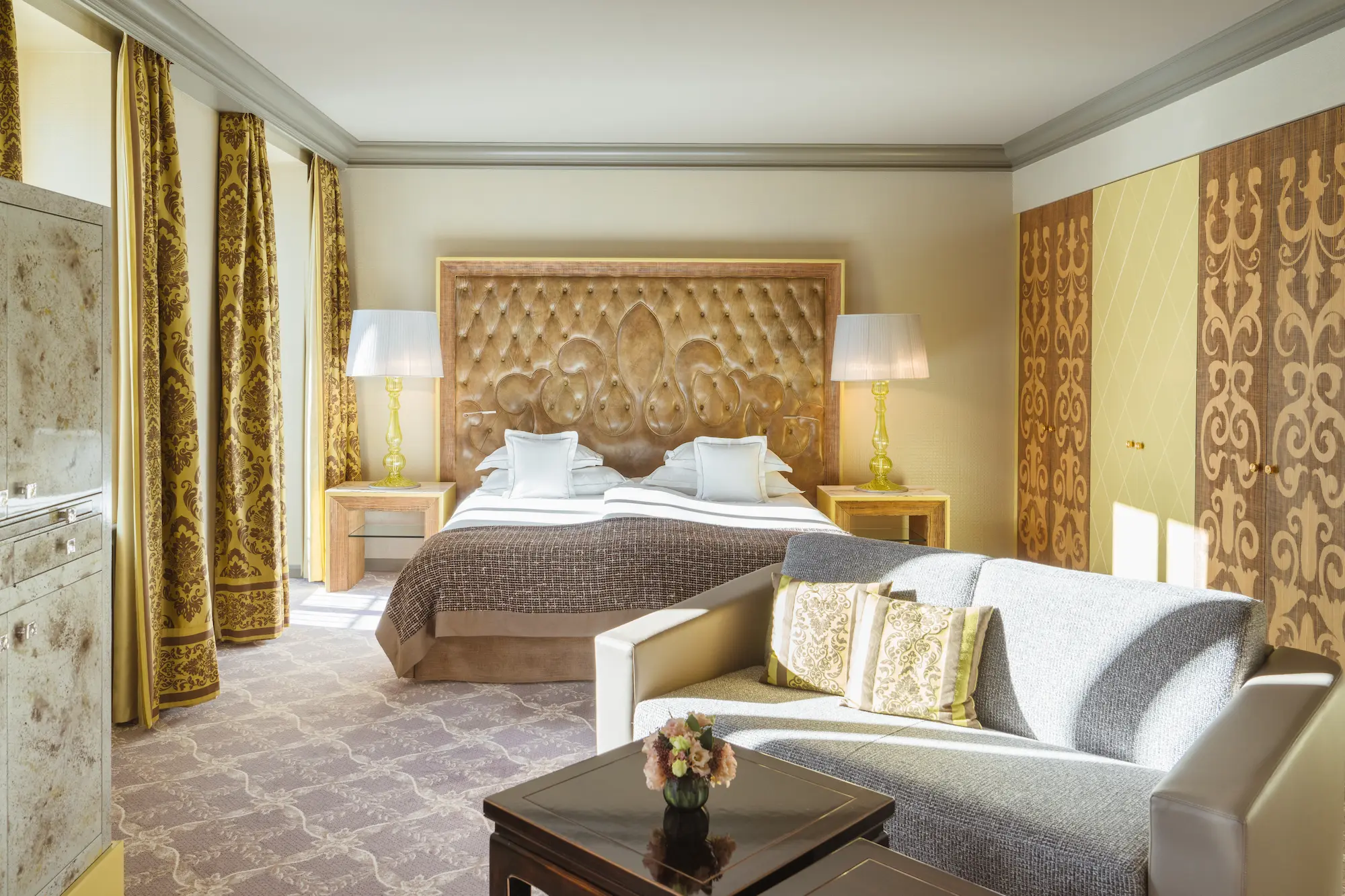 Spacious Carlton Hotel suite with ornate tufted headboard, gold and taupe decor, separate seating area, and elegant alpine-inspired furnishings
