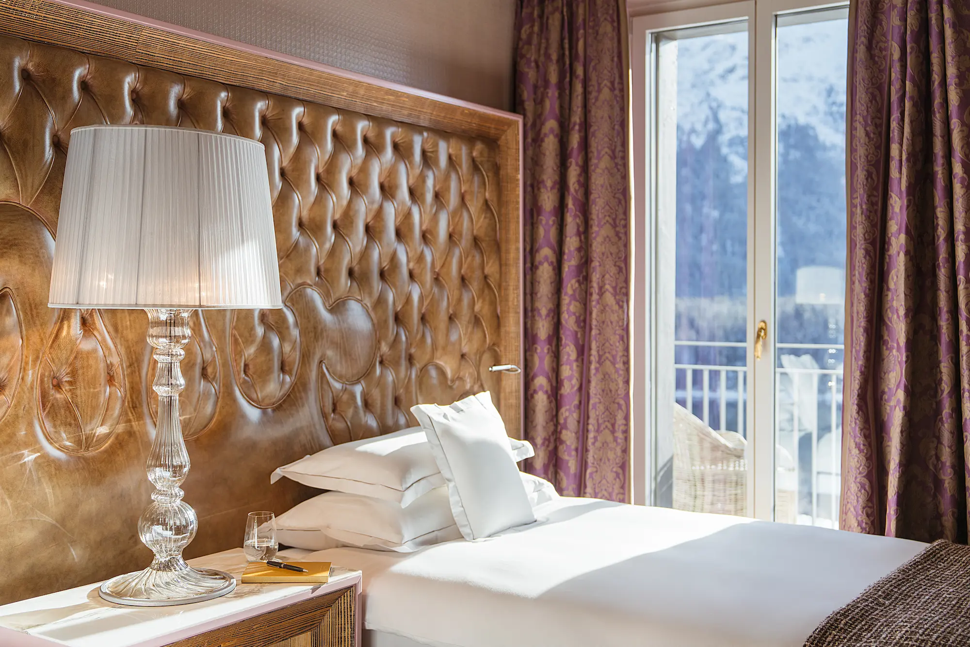 Elegant Carlton Hotel bedroom featuring tufted leather headboard, crystal table lamp, crisp white bedding, and balcony doors opening to alpine mountain vista