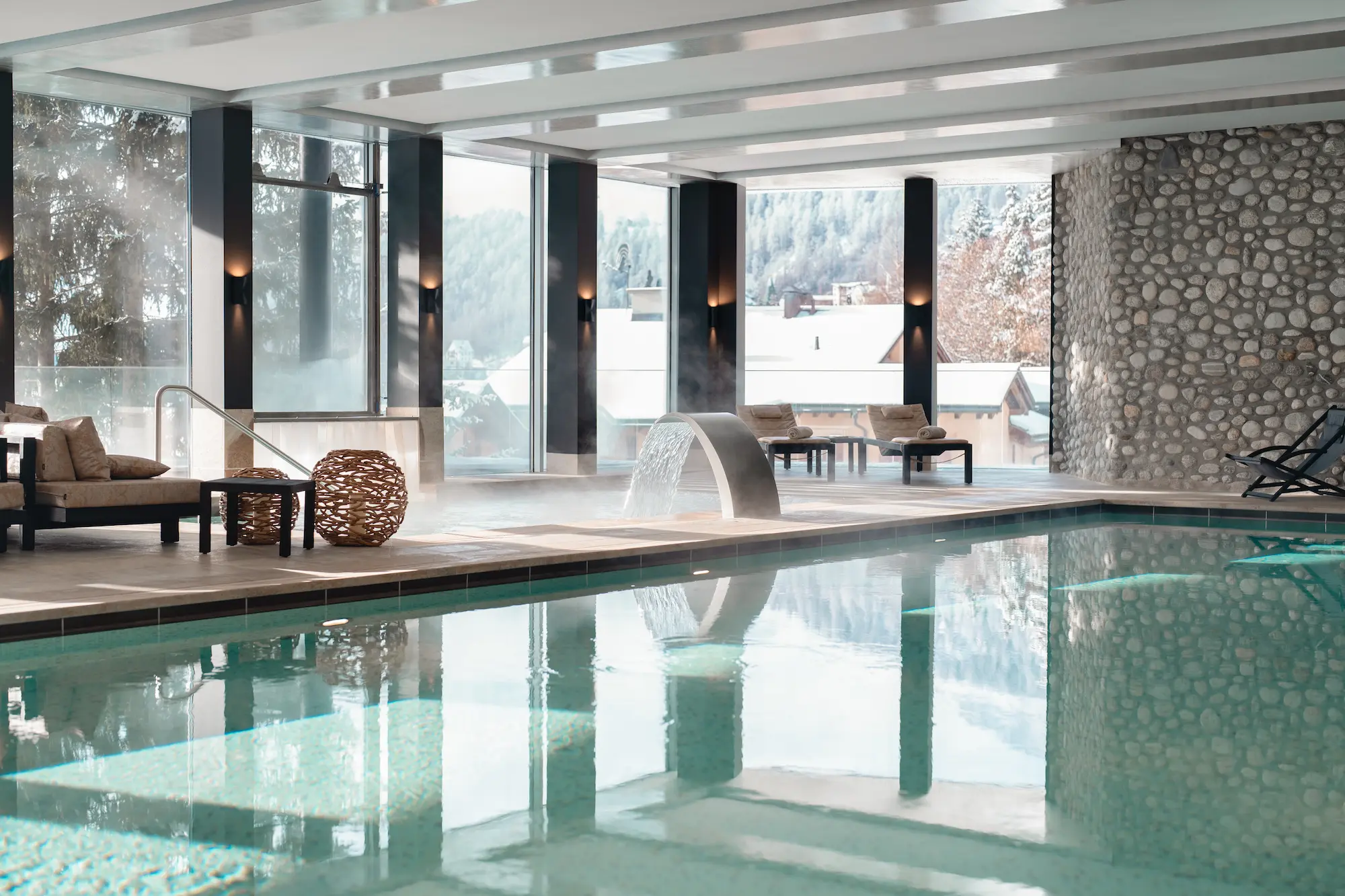 Luxurious Carlton Hotel spa featuring heated indoor pool with water feature, pebble wall, loungers, and floor-to-ceiling windows overlooking snowy mountains