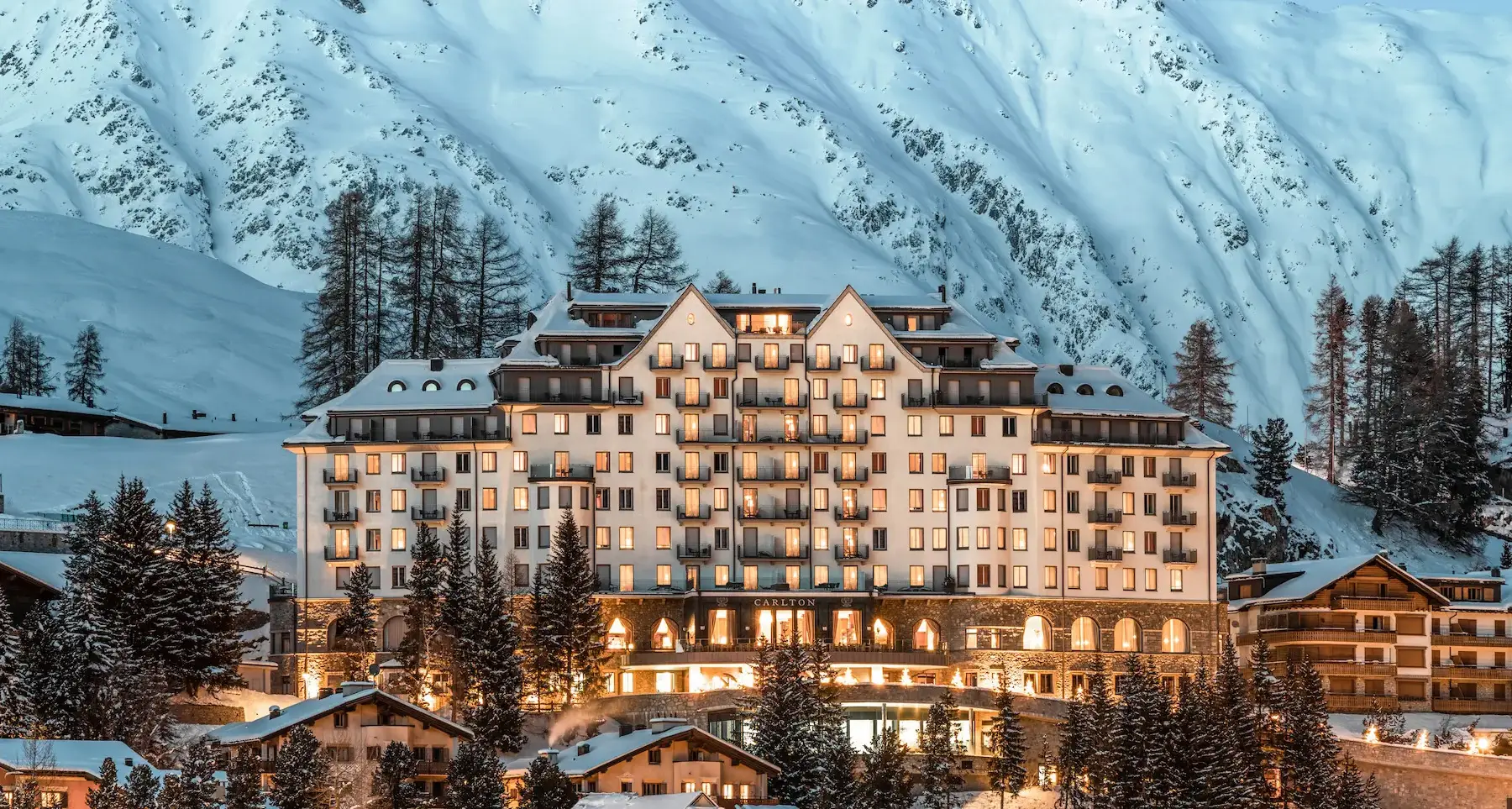 Carlton Hotel St. Moritz illuminated at dusk with dramatic snow-covered alpine mountain backdrop