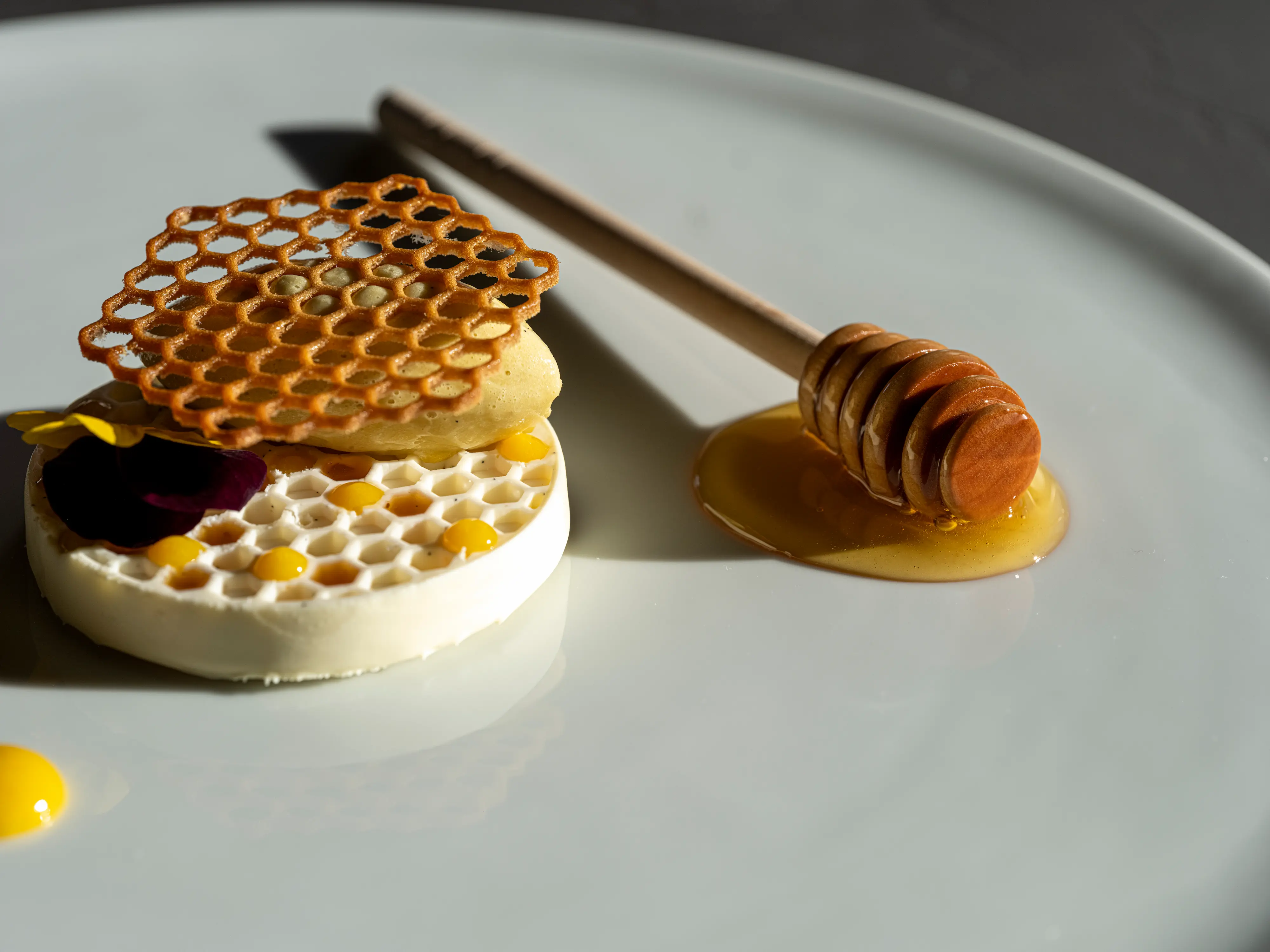 Artistic Da Vittorio dessert featuring honeycomb wafer, vanilla mousse with edible flower on white plate with honey dipper and golden honey