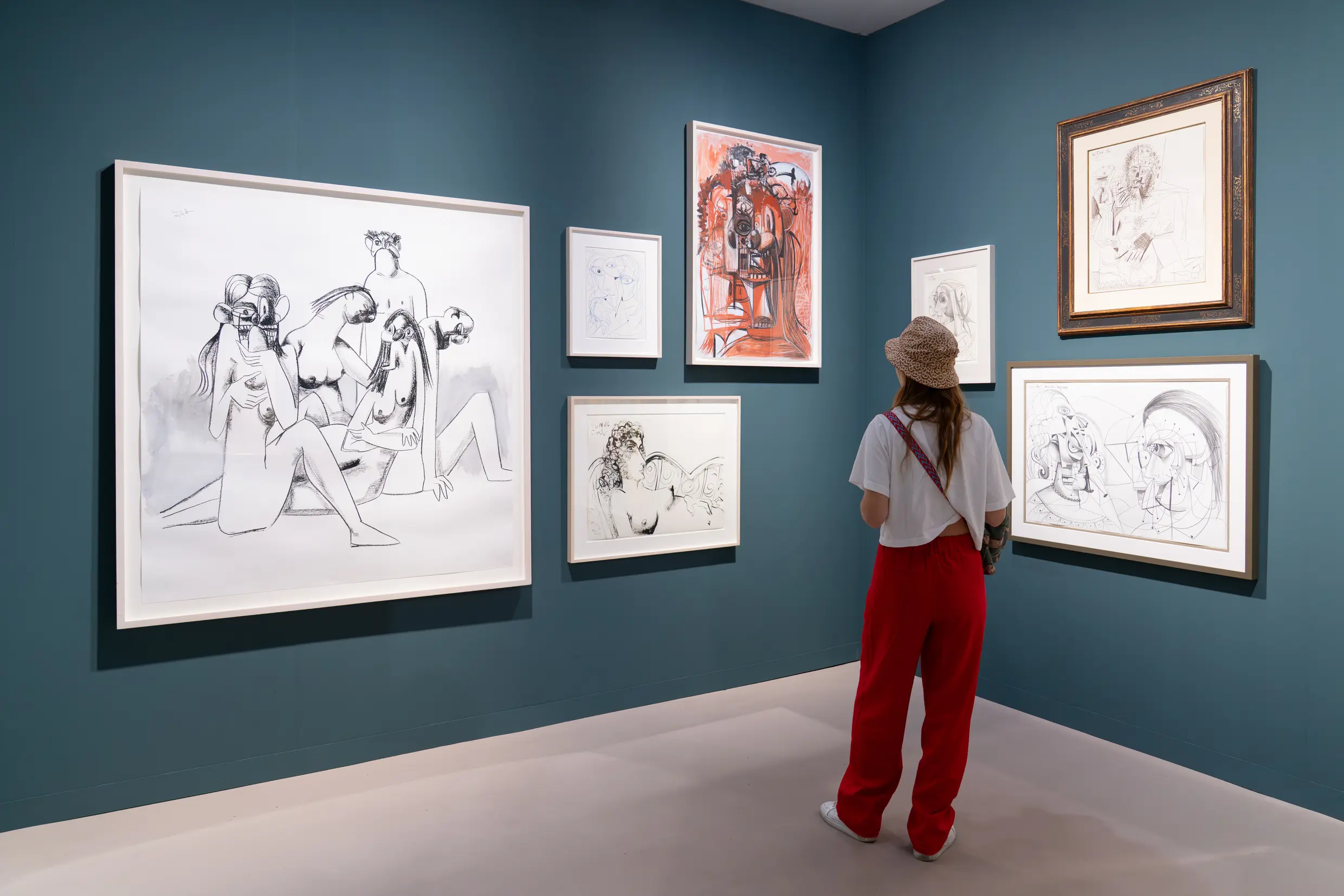 Art Basel Miami Beach 2025 gallery with teal blue walls displaying collection of figurative drawings and sketches, visitor examining contemporary artworks in curated booth