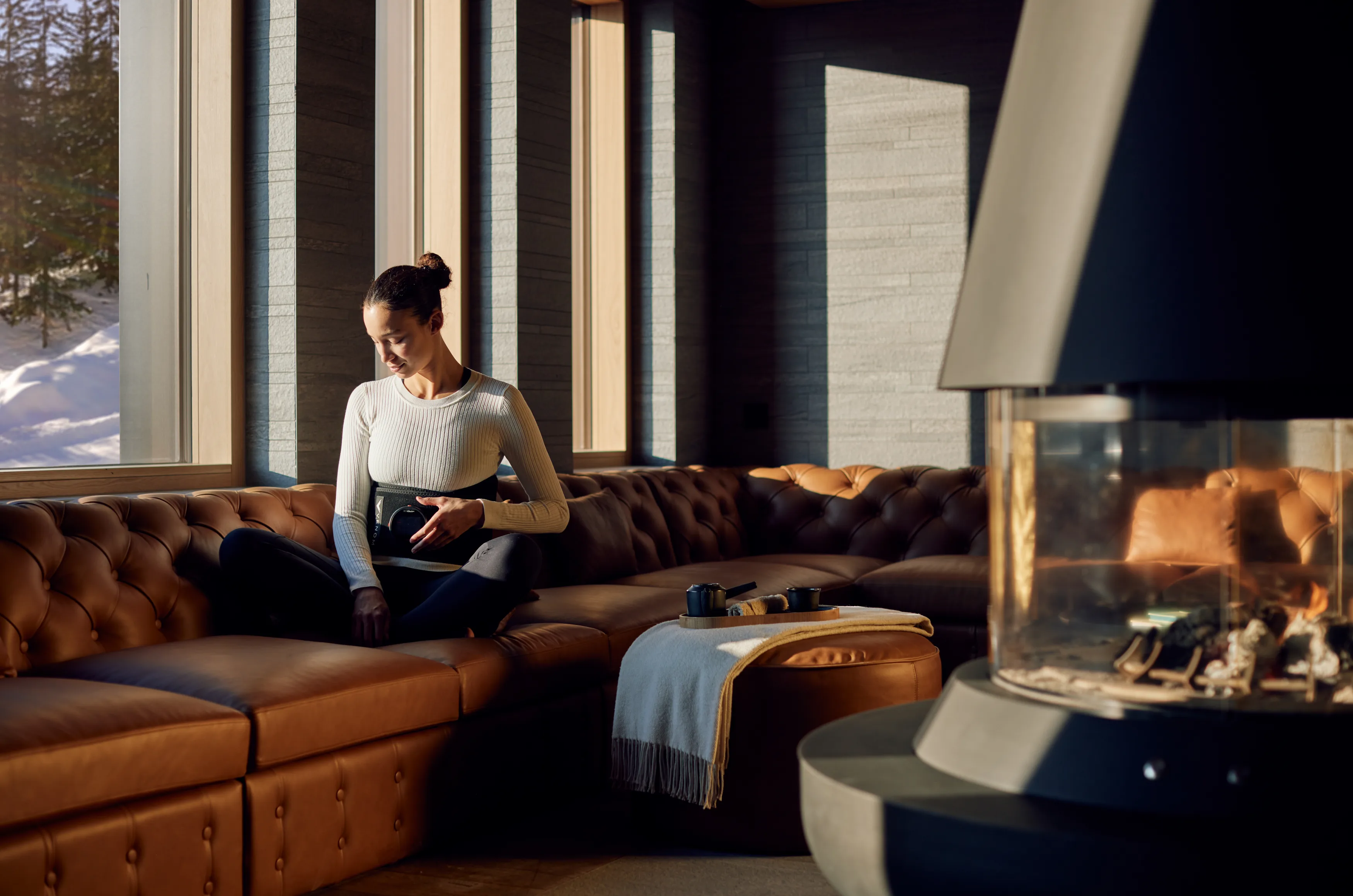 Guest relaxing in Six Senses Crans-Montana Biohack Recovery Lounge with tufted leather sofa, modern fireplace, and floor-to-ceiling windows overlooking snowy alpine landscape