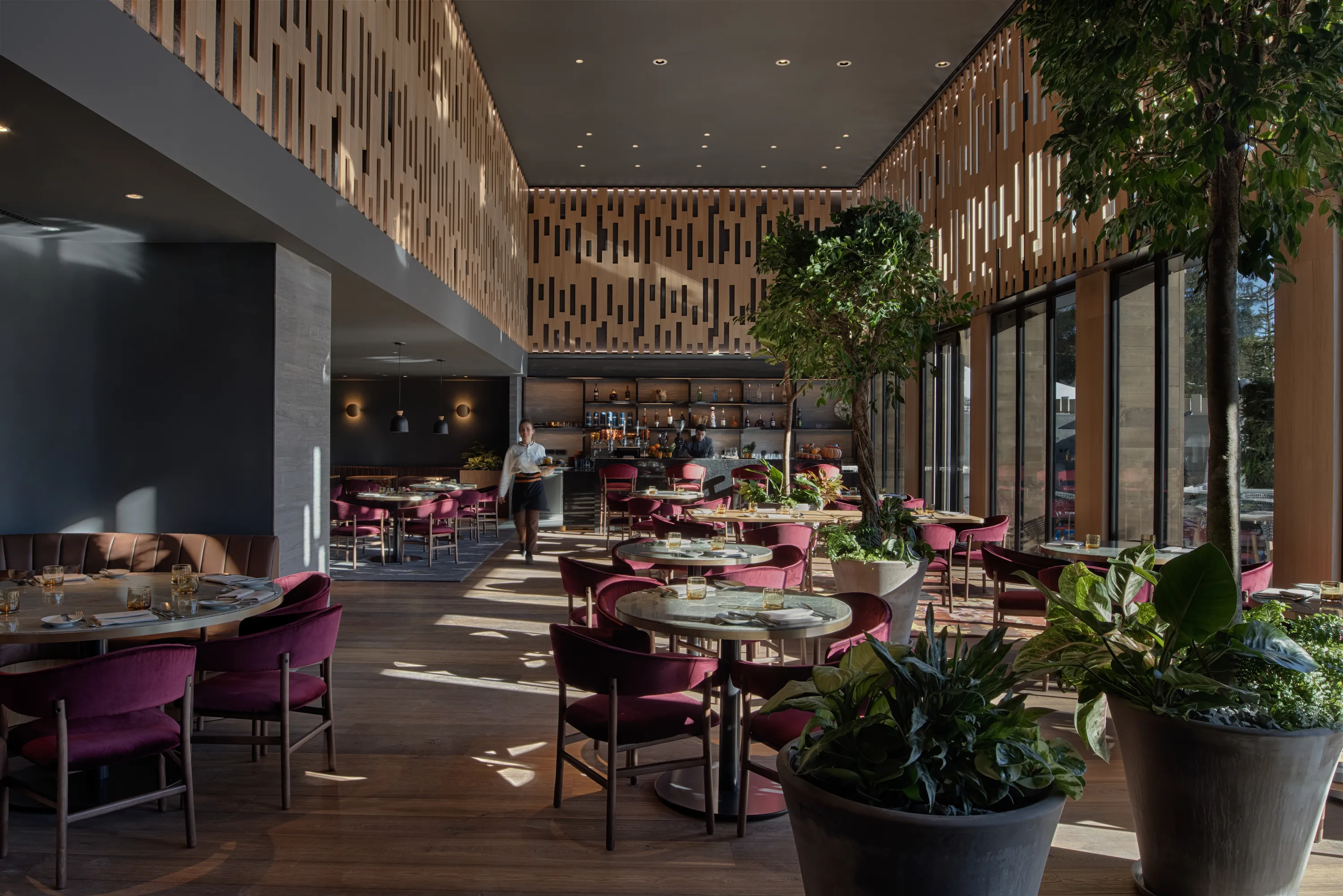Wild Cabin brasserie at Six Senses Crans-Montana with burgundy velvet seating, wooden ceiling details, abundant greenery, and natural light from floor-to-ceiling windows