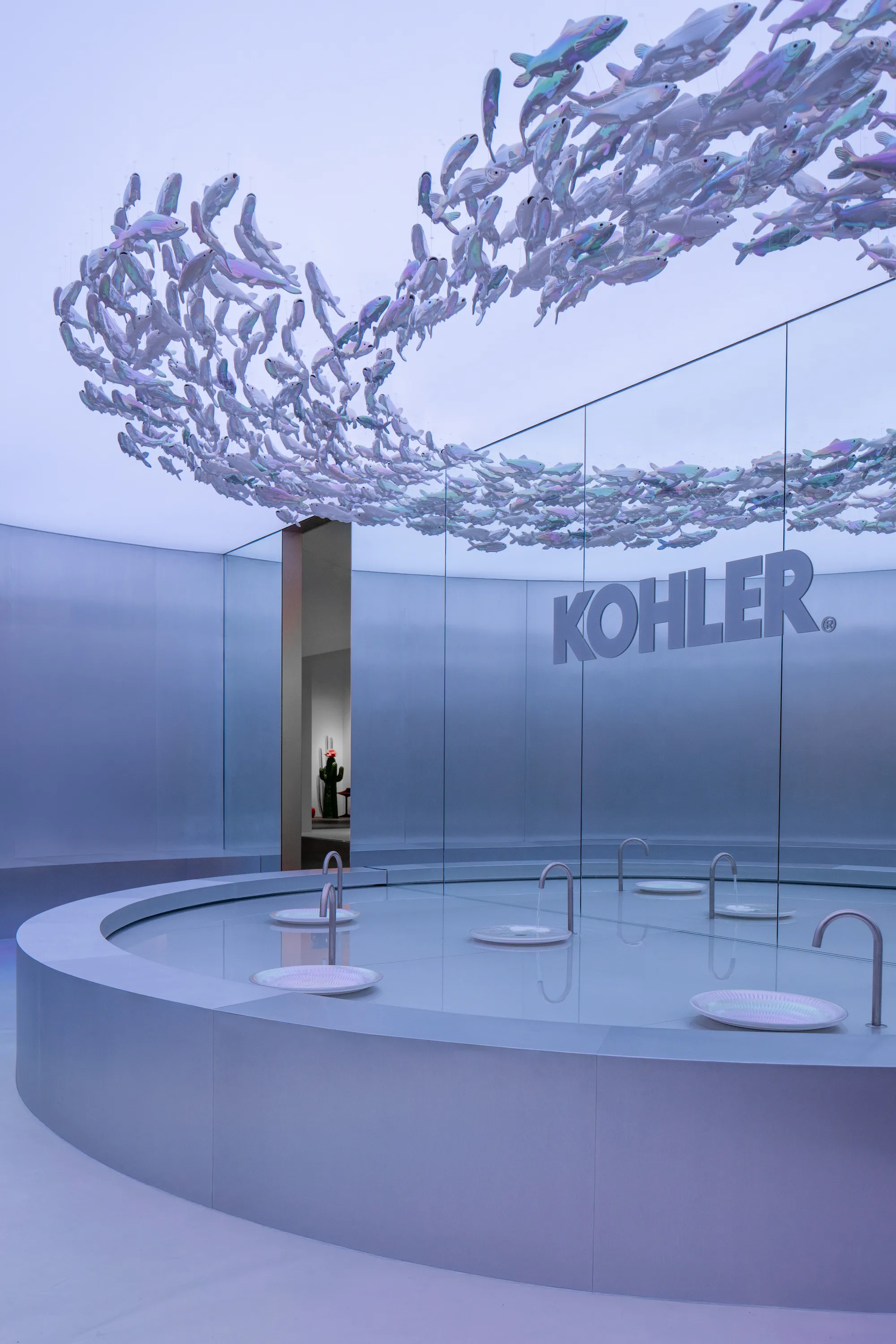 Kohler immersive installation at Design Miami 2025 featuring serene blue environment with suspended iridescent sculptural fish installation, circular display platform showcasing contemporary faucet designs and basin fixtures