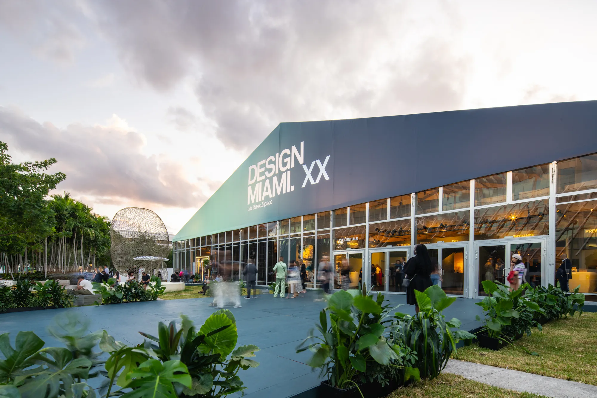 Design Miami 2025 pavilion at Pride Park featuring modern glass facade with XX anniversary branding, illuminated interior, outdoor seating areas, tropical landscaping, and visitors at dusk