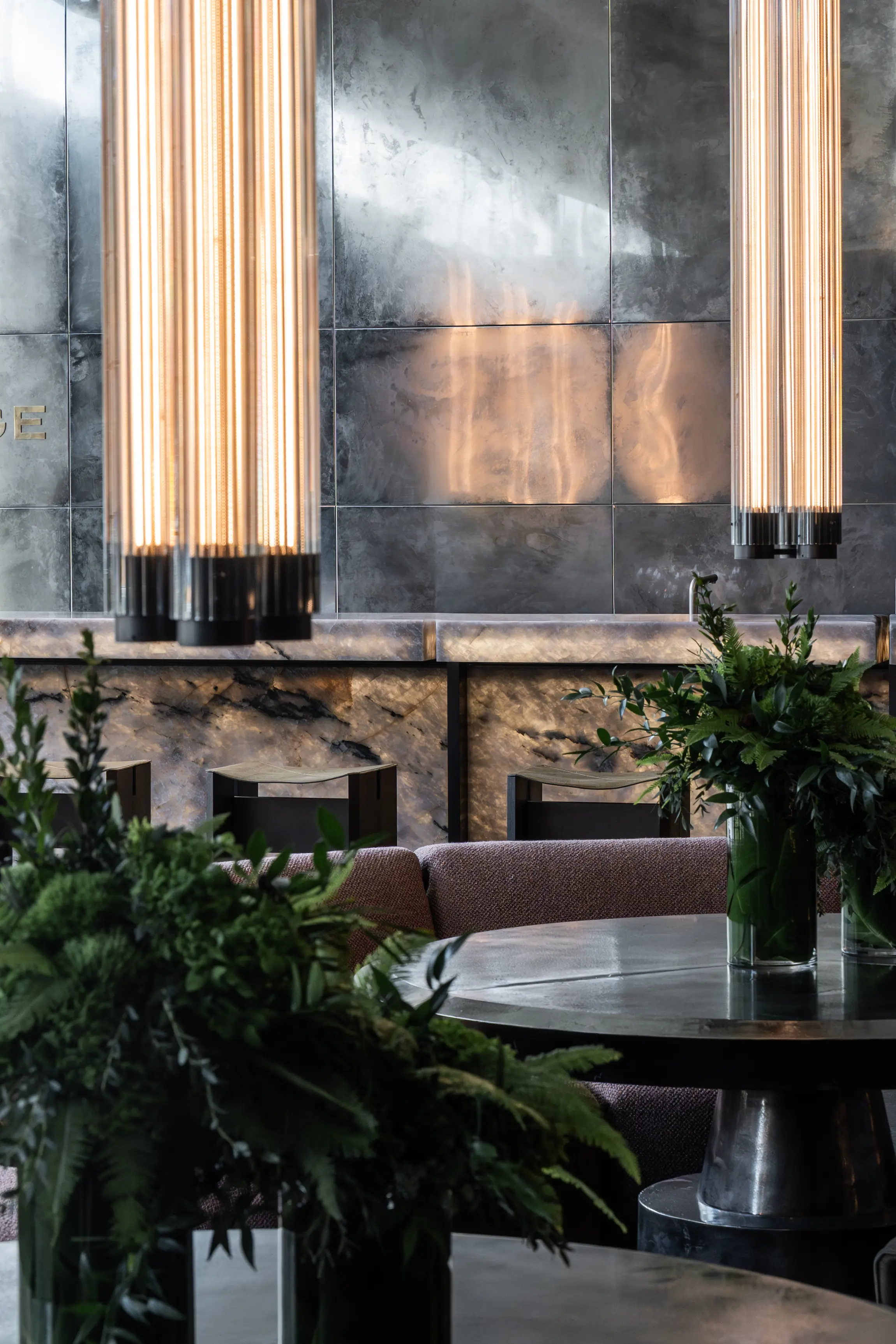 Henge Collectors Lounge detail showing vertical tubular pendant lighting against textured metal walls with sculptural stone table and greenery arrangements