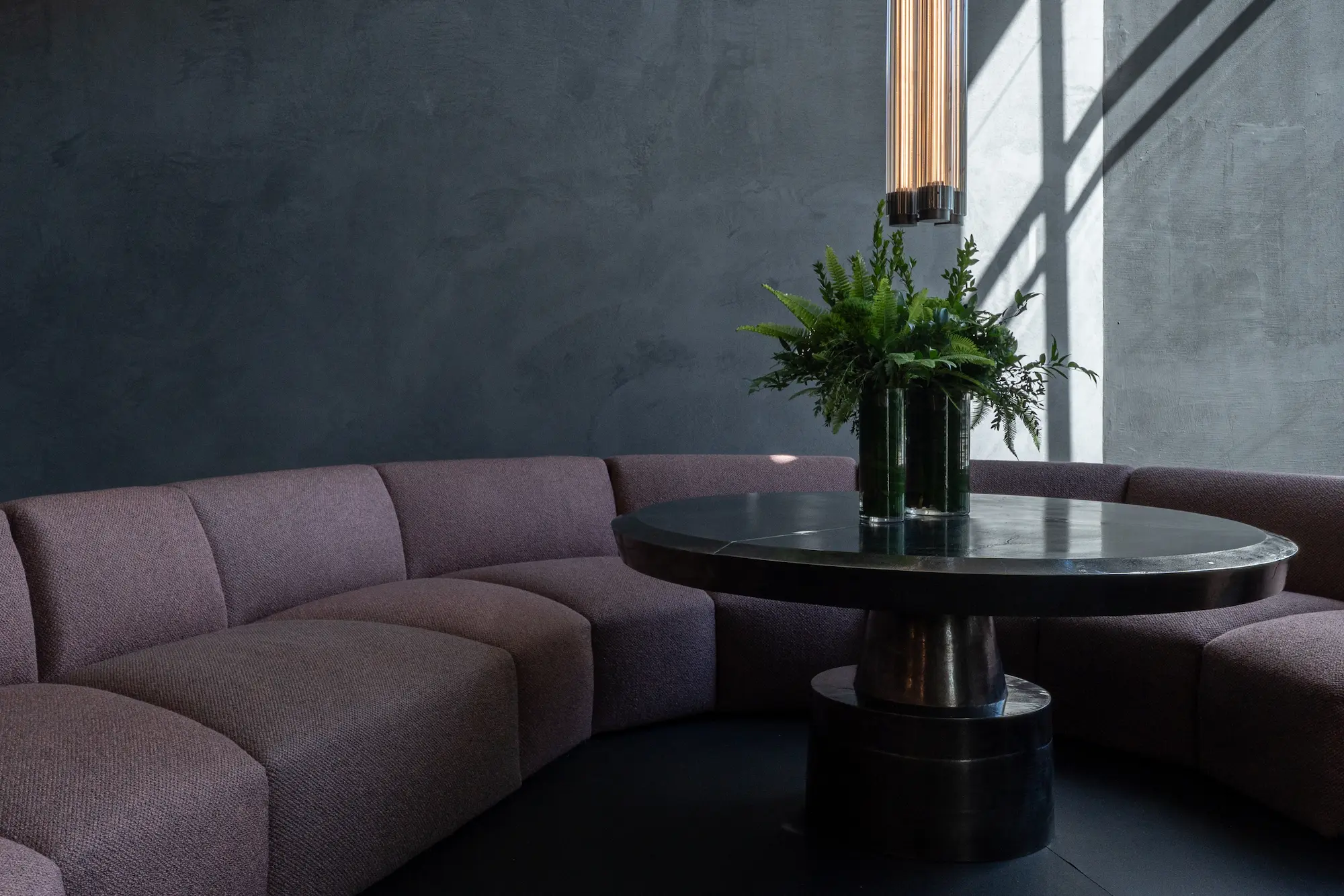 Henge Collectors Lounge featuring curved modular seating with mauve upholstery, sculptural black metal round table, vertical pendant lighting, and natural light creating dramatic shadows