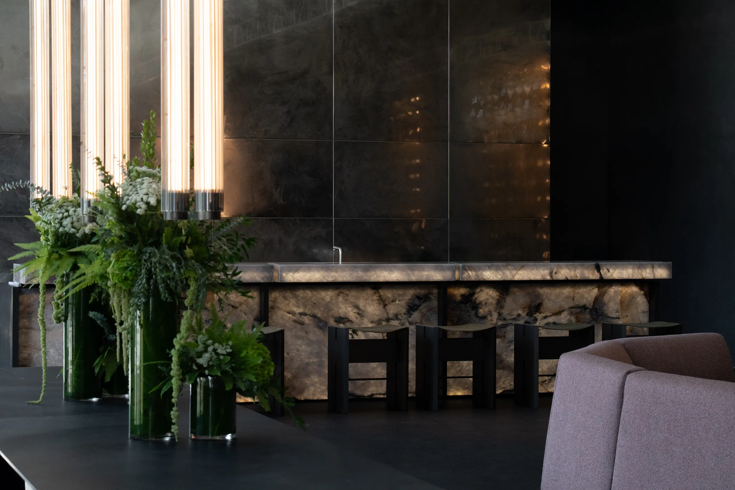 Henge Collectors Lounge detail showing vertical tubular pendant lighting against textured metal walls with sculptural stone table and greenery arrangements