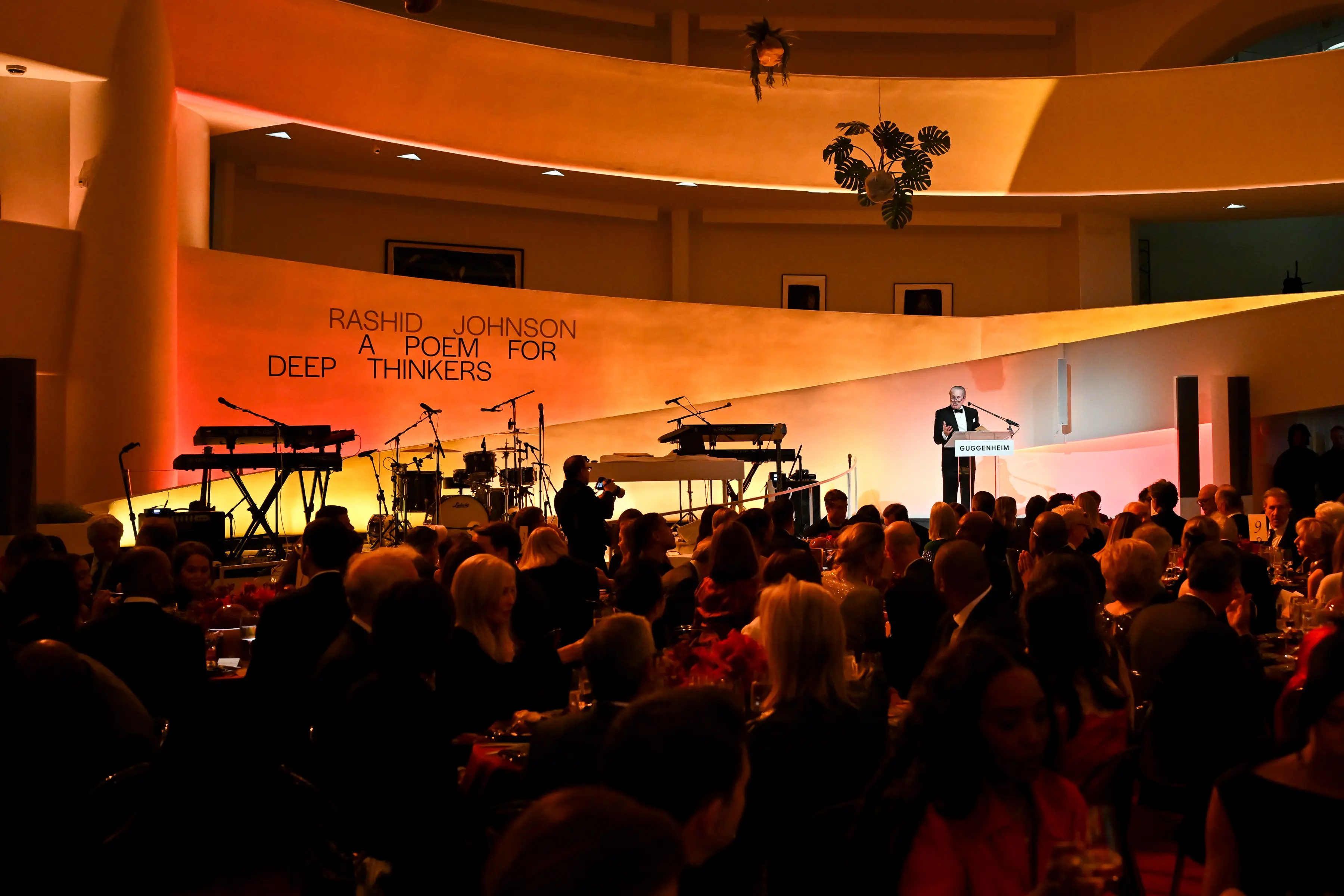 Speaker at podium during Guggenheim Museum gala celebrating Rashid Johnson exhibition A Poem for Deep Thinkers with musical instruments on stage and guests dining