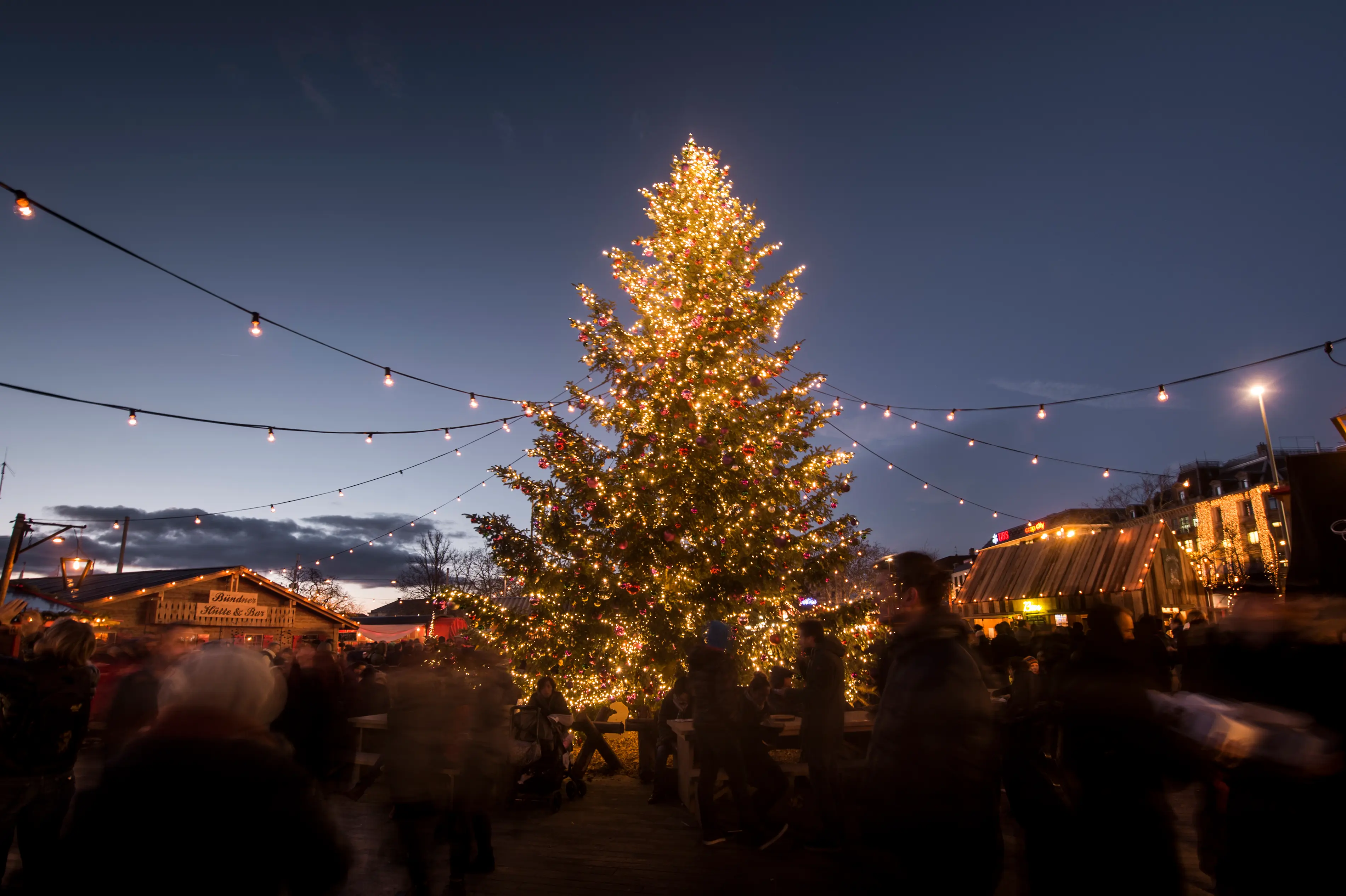 Giant illuminated Christmas tree at dusk surrounded by visitors and wooden chalet stalls at Zürcher Wienachtsdorf Christmas market on Sechseläutenplatz in Zurich, Switzerland
