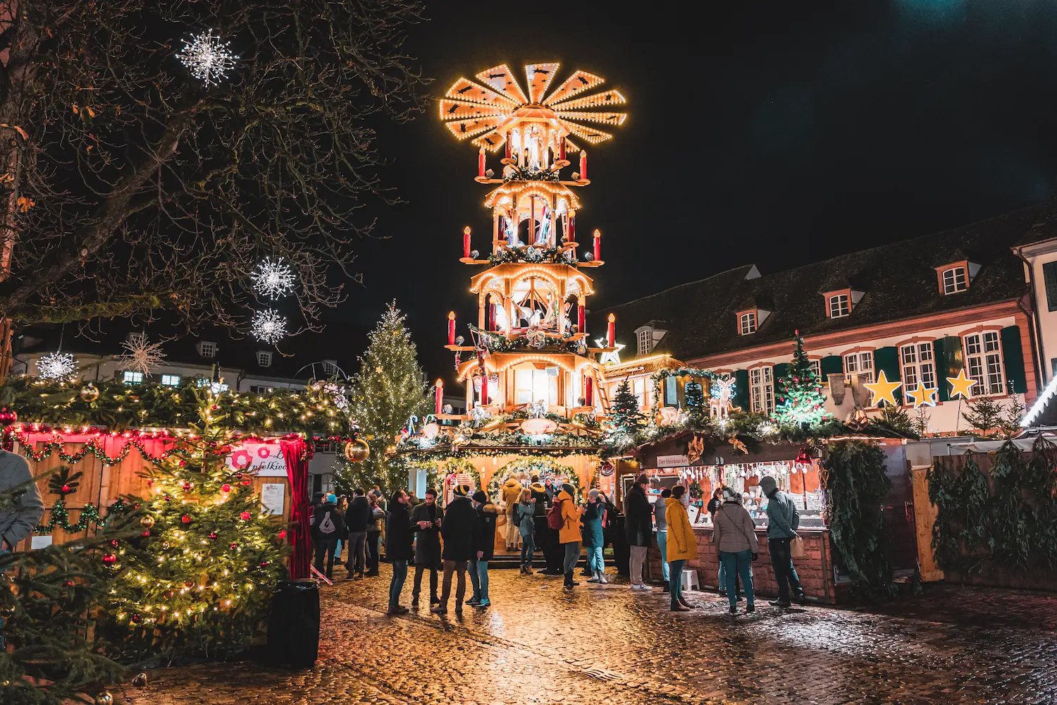 Traditional multi-tiered Weihnachtspyramide candle carousel illuminated at night surrounded by Christmas trees and wooden market stalls at Basel Christmas market near Gothic Münster cathedral