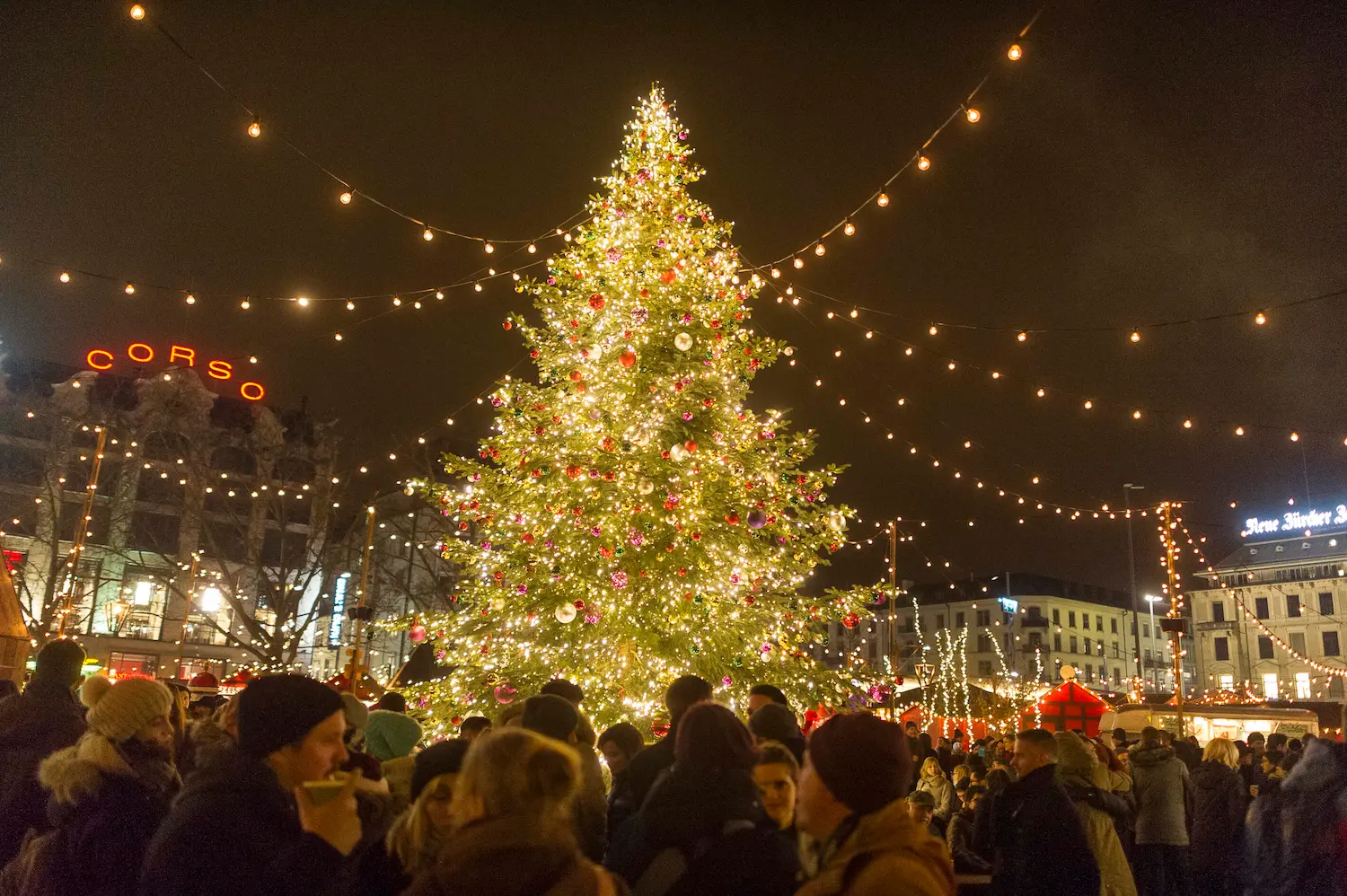 Giant illuminated Christmas tree surrounded by crowds at Zürcher Wienachtsdorf on Sechseläutenplatz with string lights and Corso sign visible in Zurich night skyline