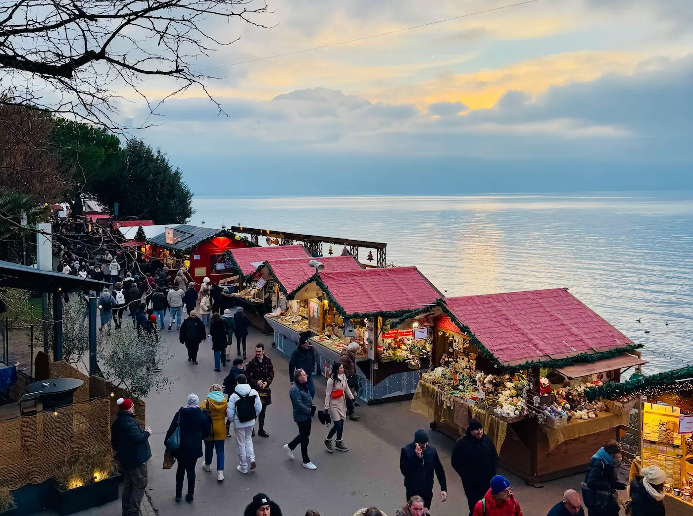 Wooden chalets with red roofs lining Lake Geneva waterfront at Montreux Noël Christmas market during sunset with visitors browsing artisan stalls and Swiss Alps in distance