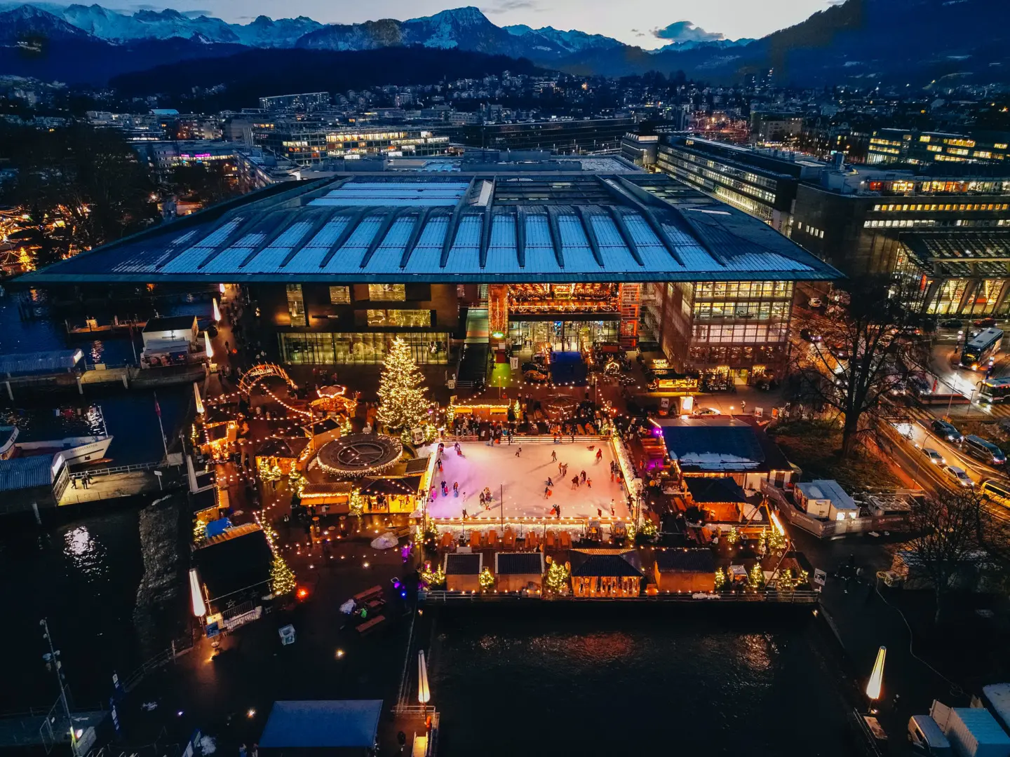 Aerial view of illuminated Eiszauber ice skating rink with Christmas tree and market chalets at dusk beside KKL Culture and Convention Centre Lucerne with Swiss Alps in background