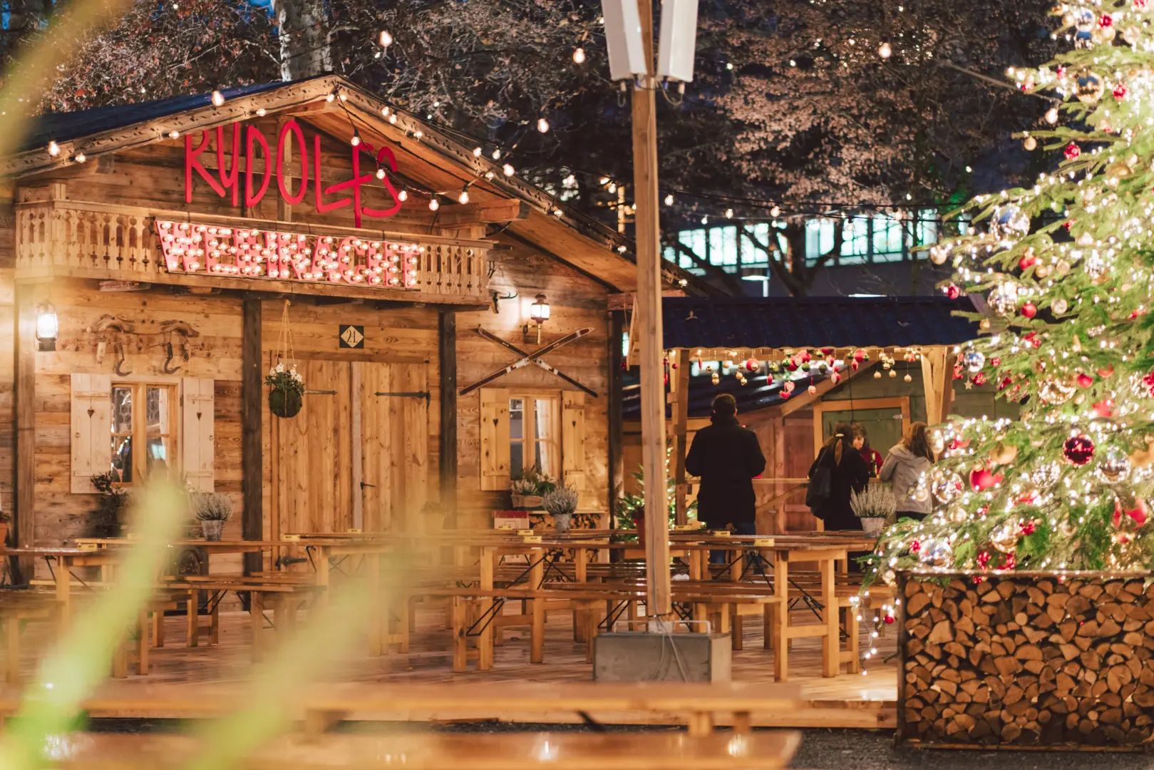Rudolfs wooden Alpine chalet restaurant with outdoor seating area, string lights, and Christmas tree at Lucerne winter market creating cozy evening atmosphere