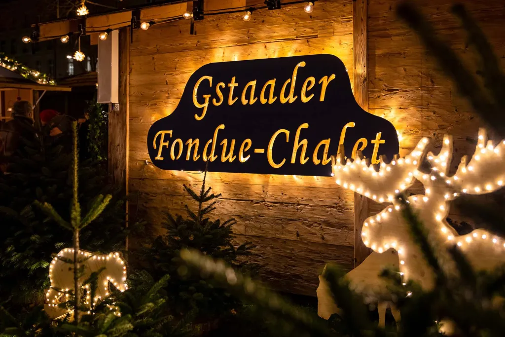 Illuminated Gstaader Fondue-Chalet sign on wooden Alpine chalet serving traditional Swiss cheese fondue at Bern Sternenmarkt Christmas market with decorative light displays