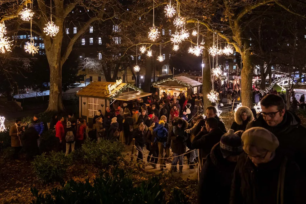 Crowds gathered at Berner Sternenmarkt Star Market with illuminated hanging star decorations in trees above wooden market huts with Swiss Parliament building visible in background at night