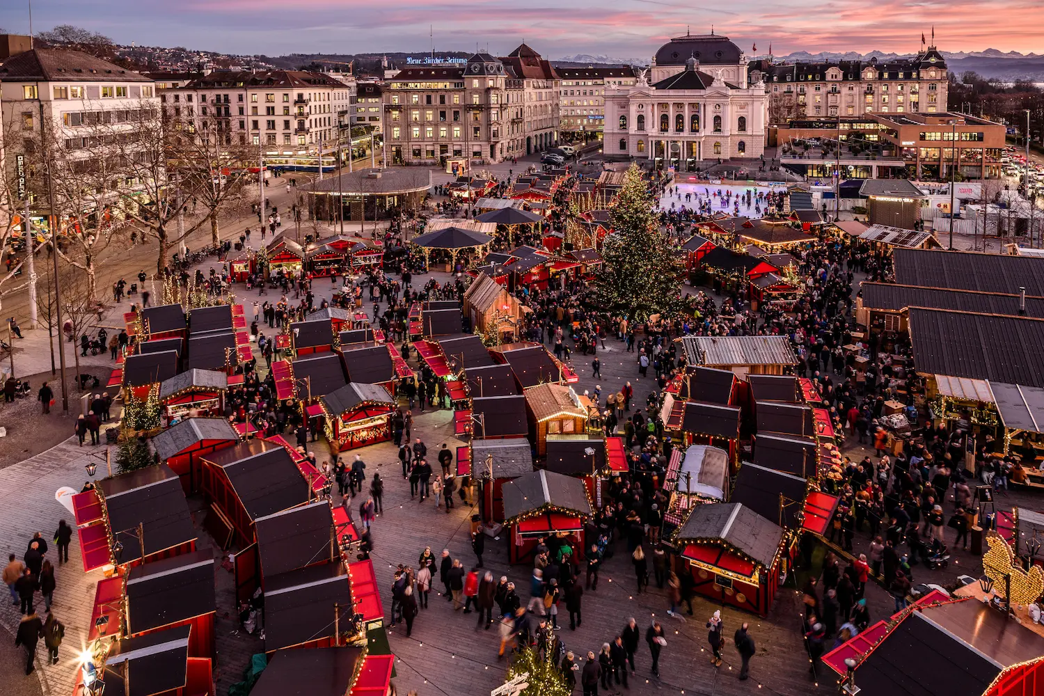 Aerial sunset view of Zürcher Wienachtsdorf Christmas market with rows of wooden chalets, ice skating rink, Christmas tree and Zurich Opera House on Sechseläutenplatz with pink sky and Alps in distance