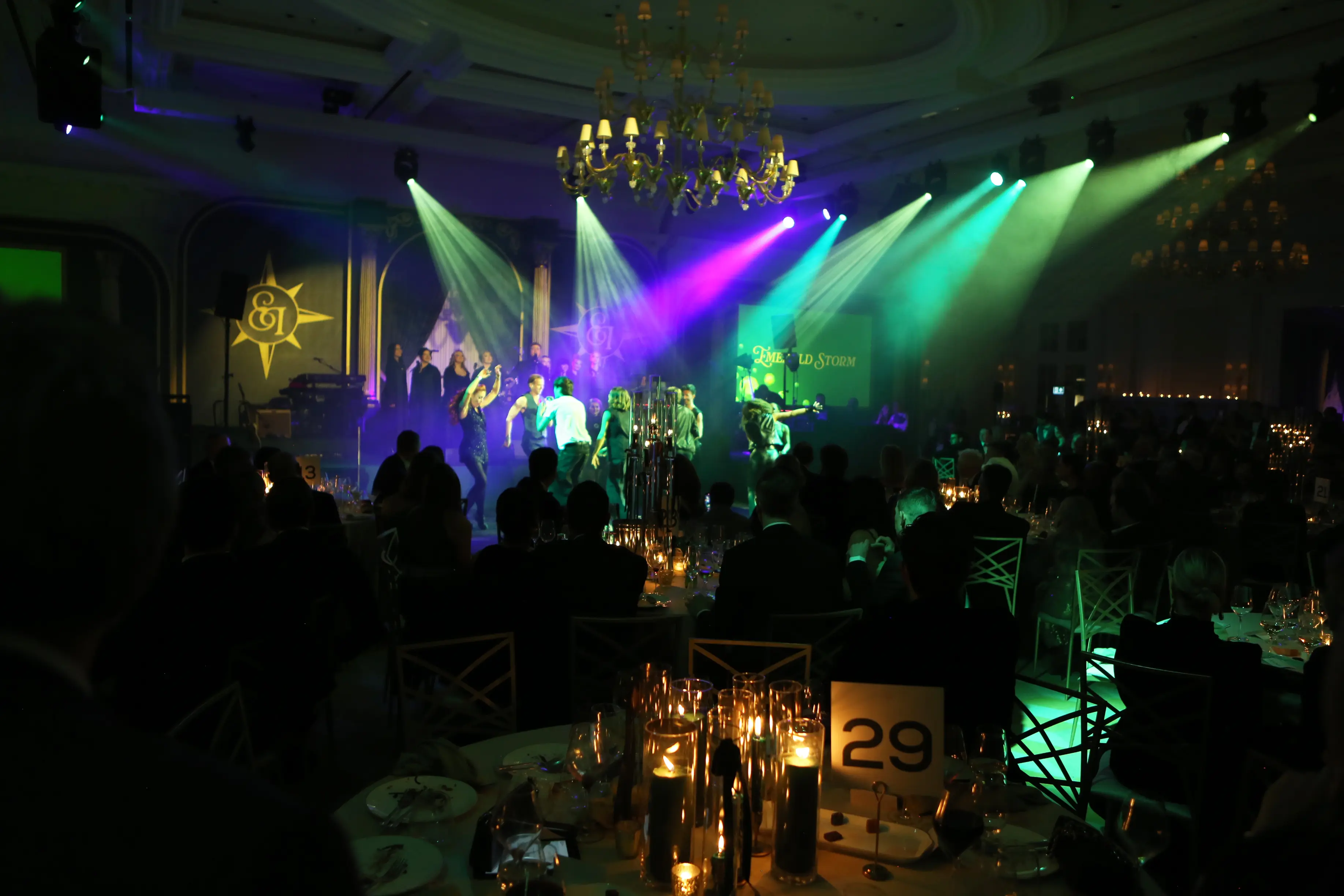 Live band performance with dramatic emerald and purple stage lighting at Emeralds & Ivy Ball 2025 charity gala at Raffles London The OWO with candlelit dining tables and guests