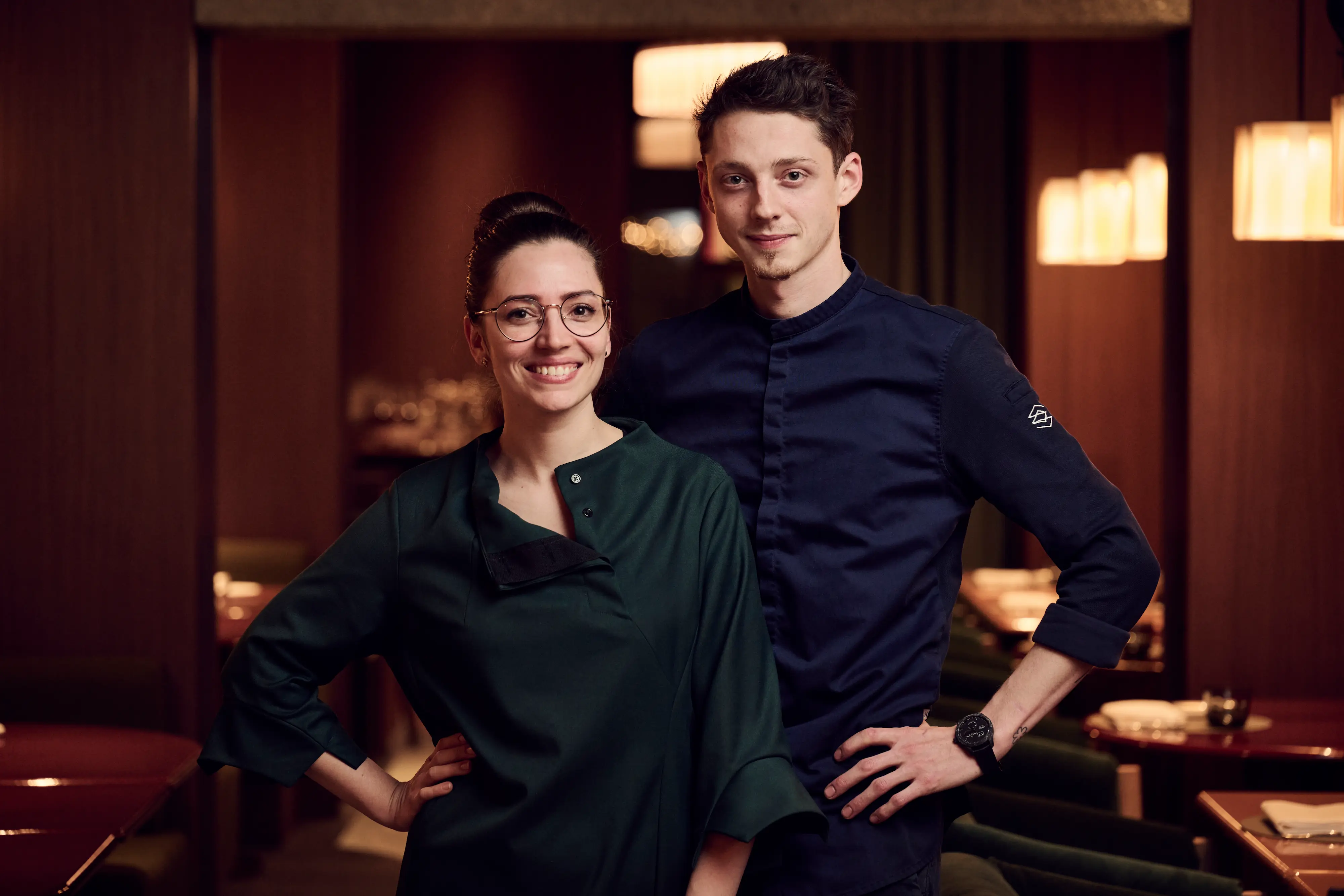 Chef Valentin Sträuli and pastry chef Hannah Vale in chef whites standing in dining room with warm lighting and emerald velvet banquettes at IGNIV Andermatt restaurant