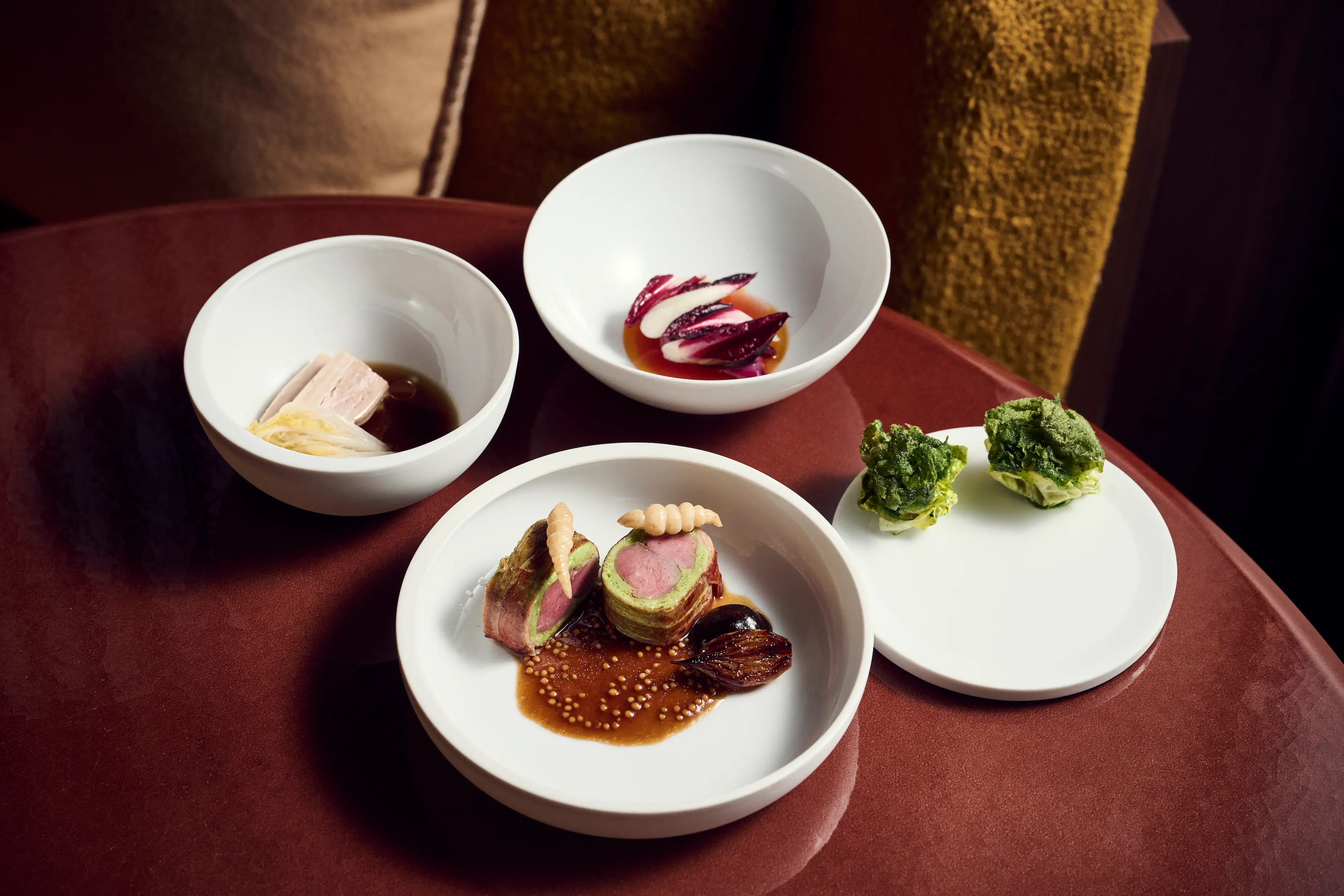 Elegant shared tasting dishes featuring duck preparation with Jerusalem artichoke and seasonal vegetables on burgundy velvet table at IGNIV Andermatt by Chef Valentin Sträuli