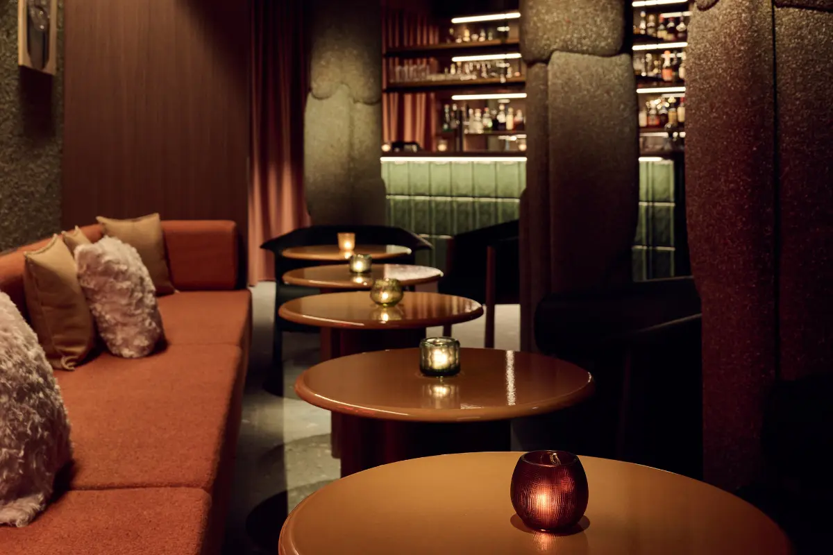 Cozy lounge area with orange velvet banquettes, round candlelit tables, and illuminated emerald green bar in background featuring Patricia Urquiola's retro 1970s-inspired interior design at IGNIV Andermatt