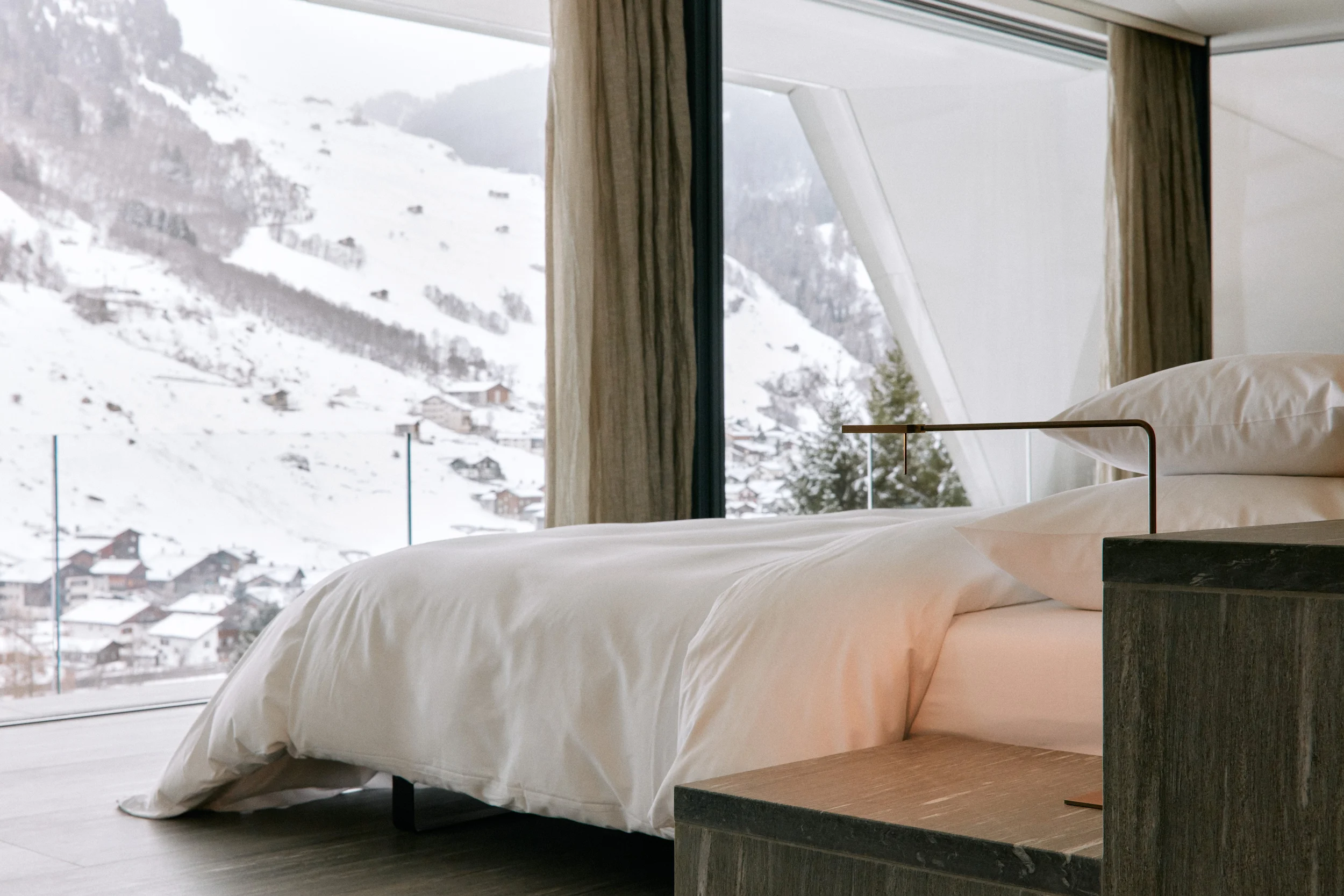 Minimalist luxury suite with floor-to-ceiling windows overlooking snow-covered Vals village and Graubünden Alps at 7132 Hotel designed by Pritzker Prize-winning architects with natural materials and featherbed