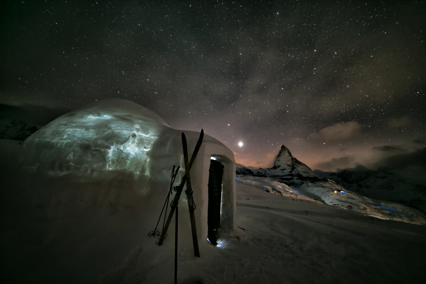 Snow igloo with skis under Milky Way galaxy and Matterhorn mountain peak for stargazing at Swiss Alps ice hotel