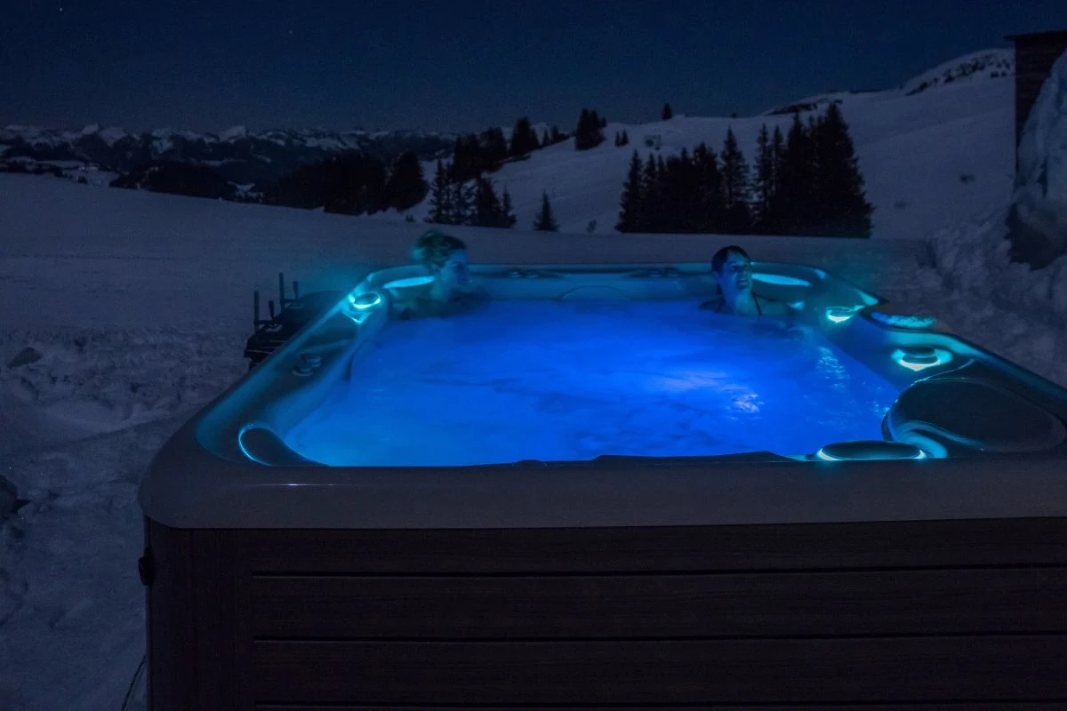 Guests relaxing in illuminated outdoor hot tub at twilight with snow-covered alpine peaks at Igloo Dorf Gstaad luxury spa experience