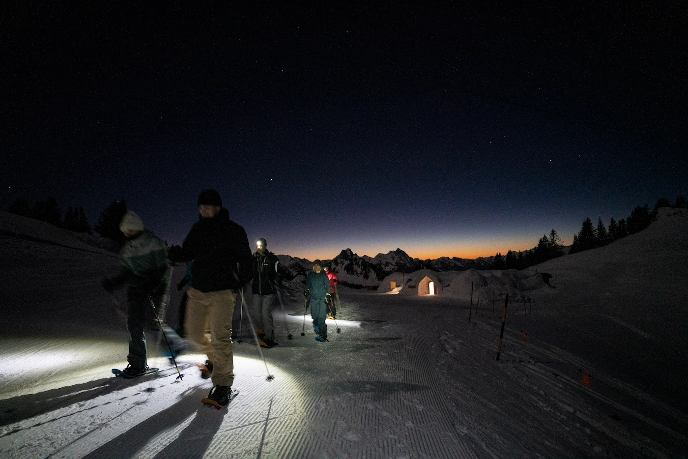 Guided nighttime snowshoe hiking excursion approaching illuminated igloo village under starlit sky at Igloo Dorf Gstaad winter adventure