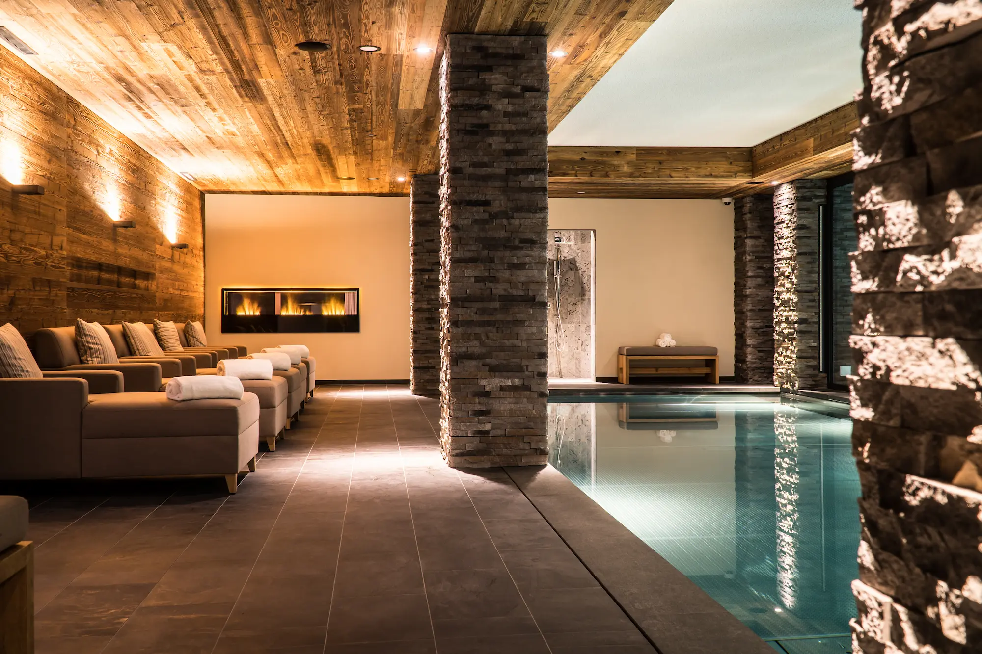 Luxury indoor hydrotherapy pool with stone columns alpine wood ceiling and fireplace lounge at The Capra Peak Health Spa Saas-Fee
