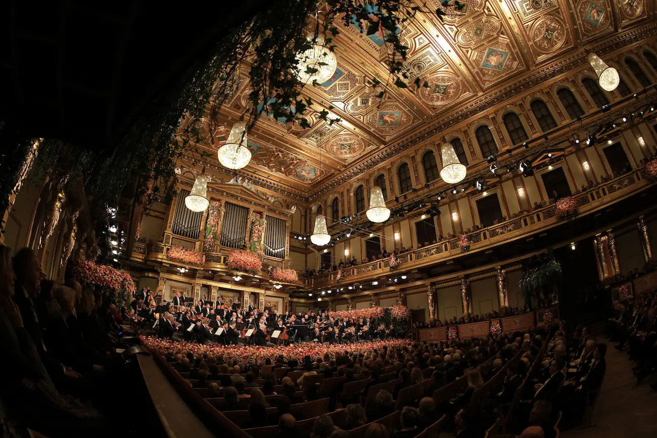 Wide-angle view of packed Golden Hall Musikverein during Vienna New Year's Concert showing gilded coffered ceiling, crystal chandeliers, multi-tiered balconies, and black-tie audience