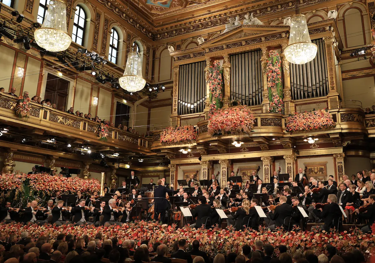 Vienna Philharmonic performing in Golden Hall Musikverein with lavish pink floral arrangements, crystal chandeliers, baroque organ, and elegant audience at New Year's Concert 2026