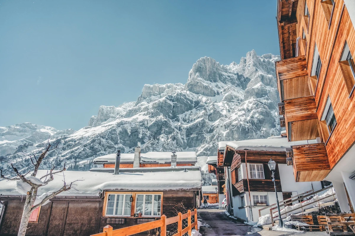Traditional wooden chalets with snow-covered roofs in Leukerbad village, dramatic Gemmi Pass rock formations and snow-covered peaks towering above, clear blue winter sky, authentic Alpine architecture at 1,400m altitude