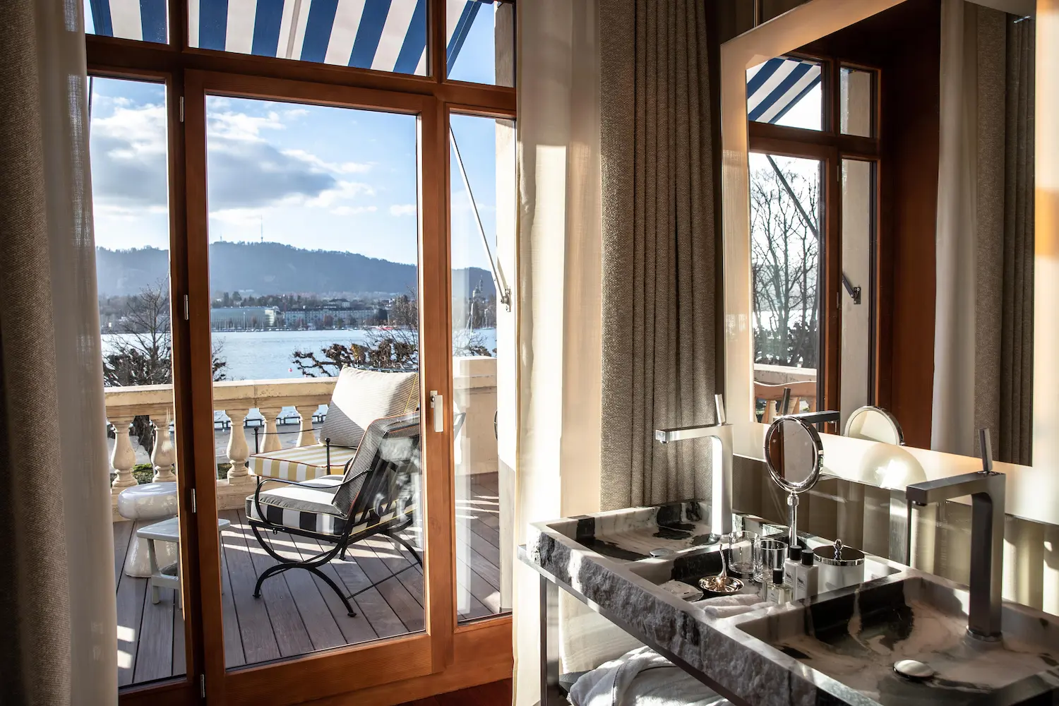 Elegant marble bathroom in Premier Terrace Room with floor-to-ceiling windows, private terrace overlooking winter Lake Zurich, Philippe Starck design, blue-and-white striped awnings visible outside