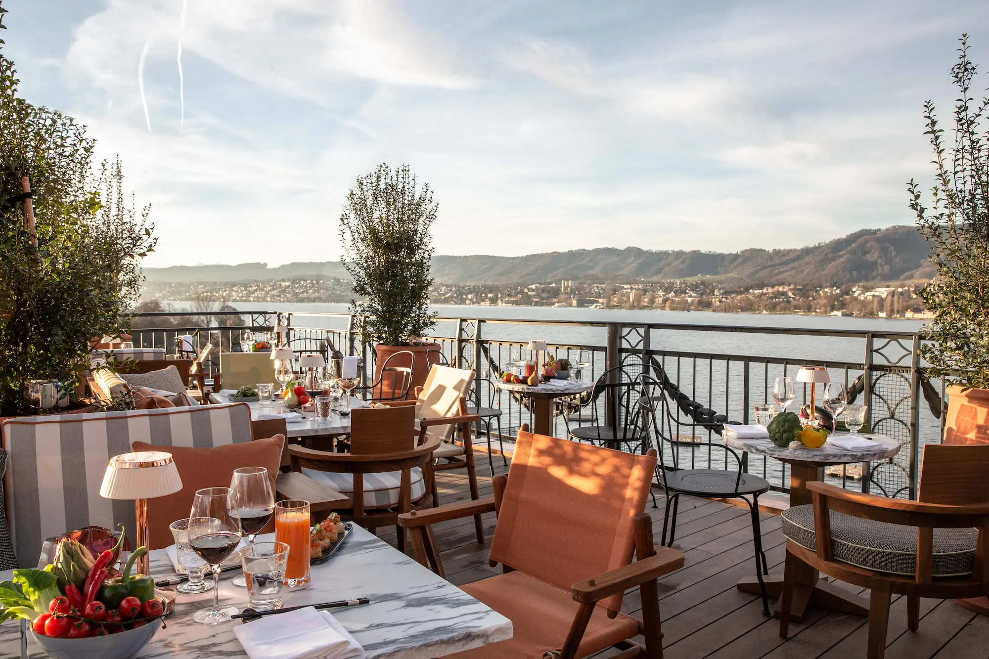 Outdoor dining terrace at La Muña rooftop restaurant with set tables, orange designer chairs, panoramic Lake Zurich views, tree-lined deck, and Alpine backdrop at La Réserve Eden au Lac