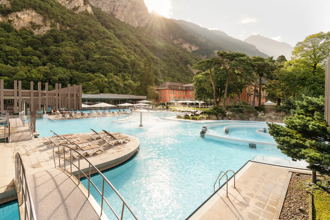 Expansive outdoor thermal pool complex at Les Bains de Lavey with turquoise 65°C waters, sun loungers, central geysers, Grand Hôtel des Bains, and lush Alpine mountains with morning sunlight in Valais