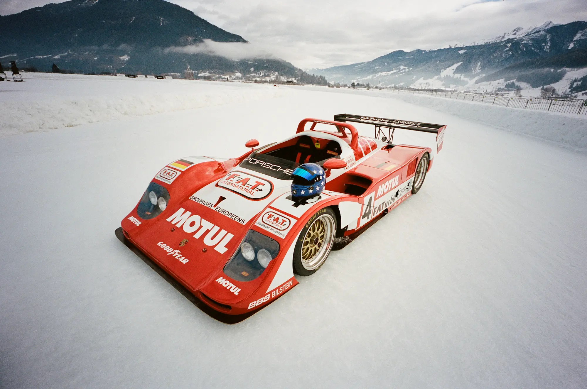 Historic red FAT Porsche 962 Le Mans race car with Motul livery on frozen runway at Zell am See Airport, snow-covered Austrian Alps in background, FAT Ice Race 2026
