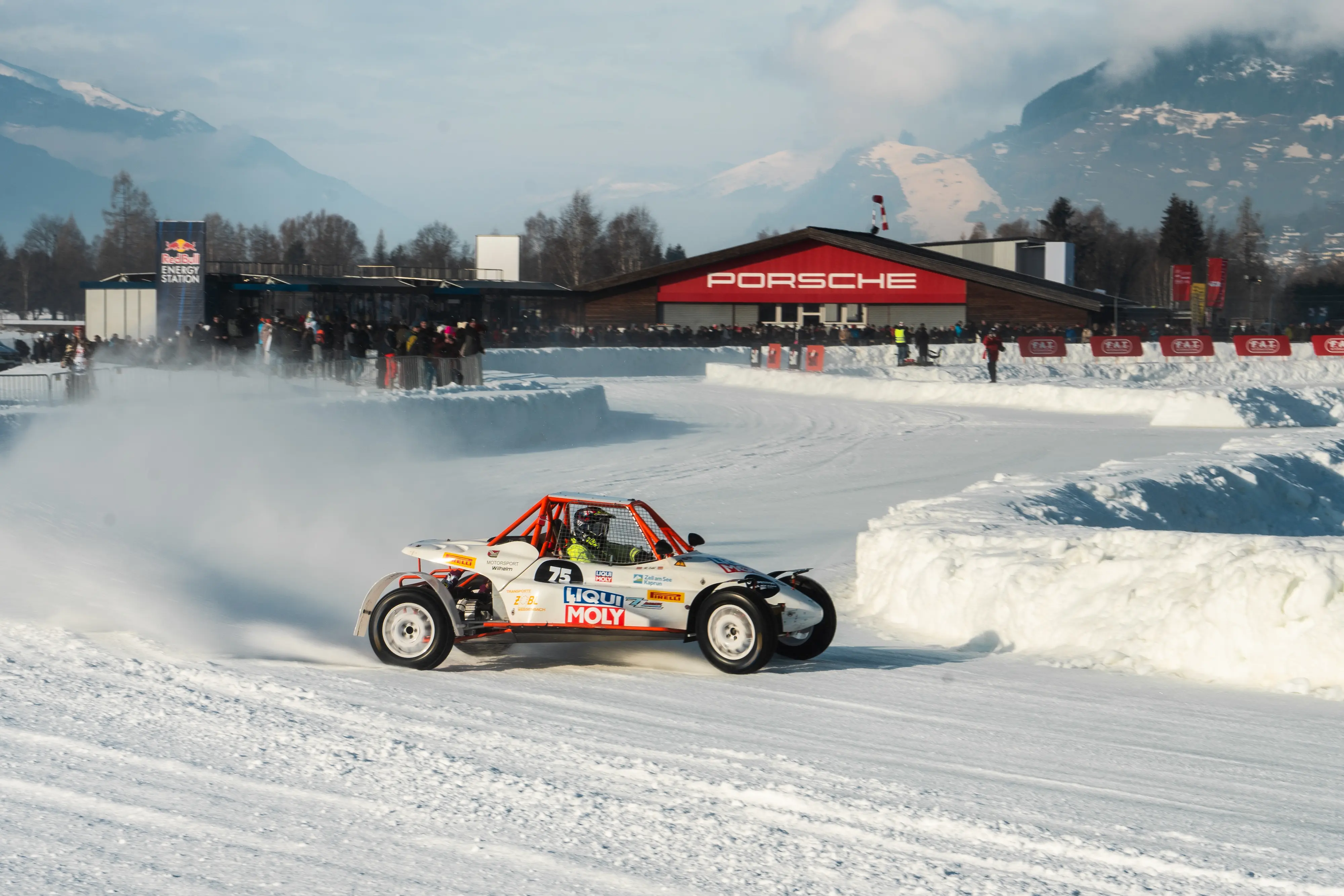 White and orange Liqui Moly sponsored ice buggy number 73 drifting with snow spray at FAT Ice Race, Porsche hangar and Red Bull station visible with Austrian Alps backdrop